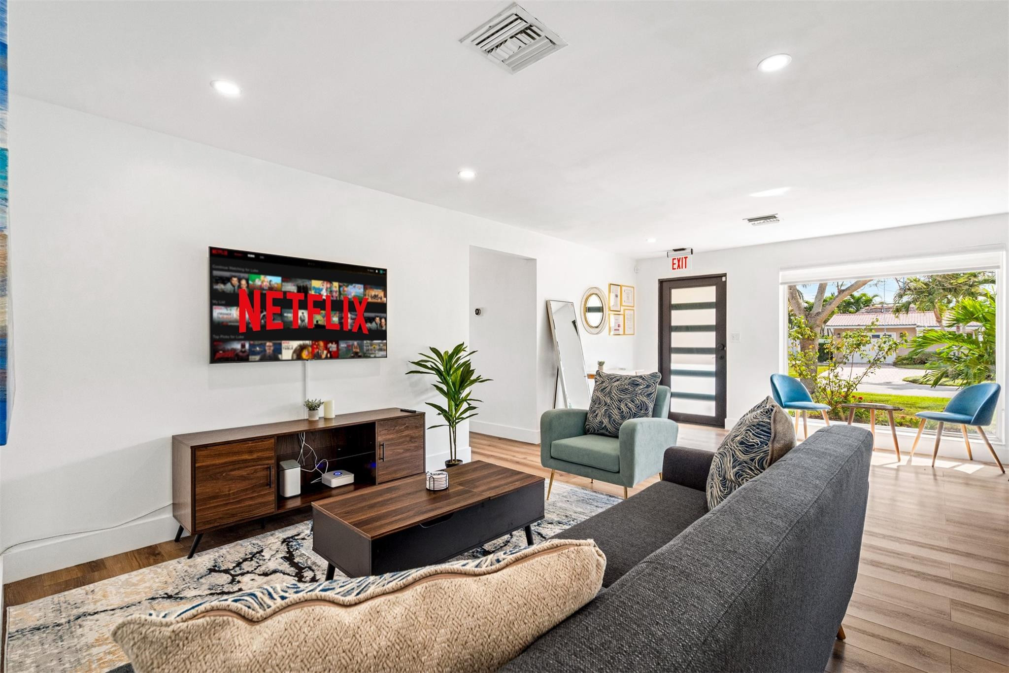 Furnished living room with gray sofa, teal armchair, and large window overlooking a leafy yard.