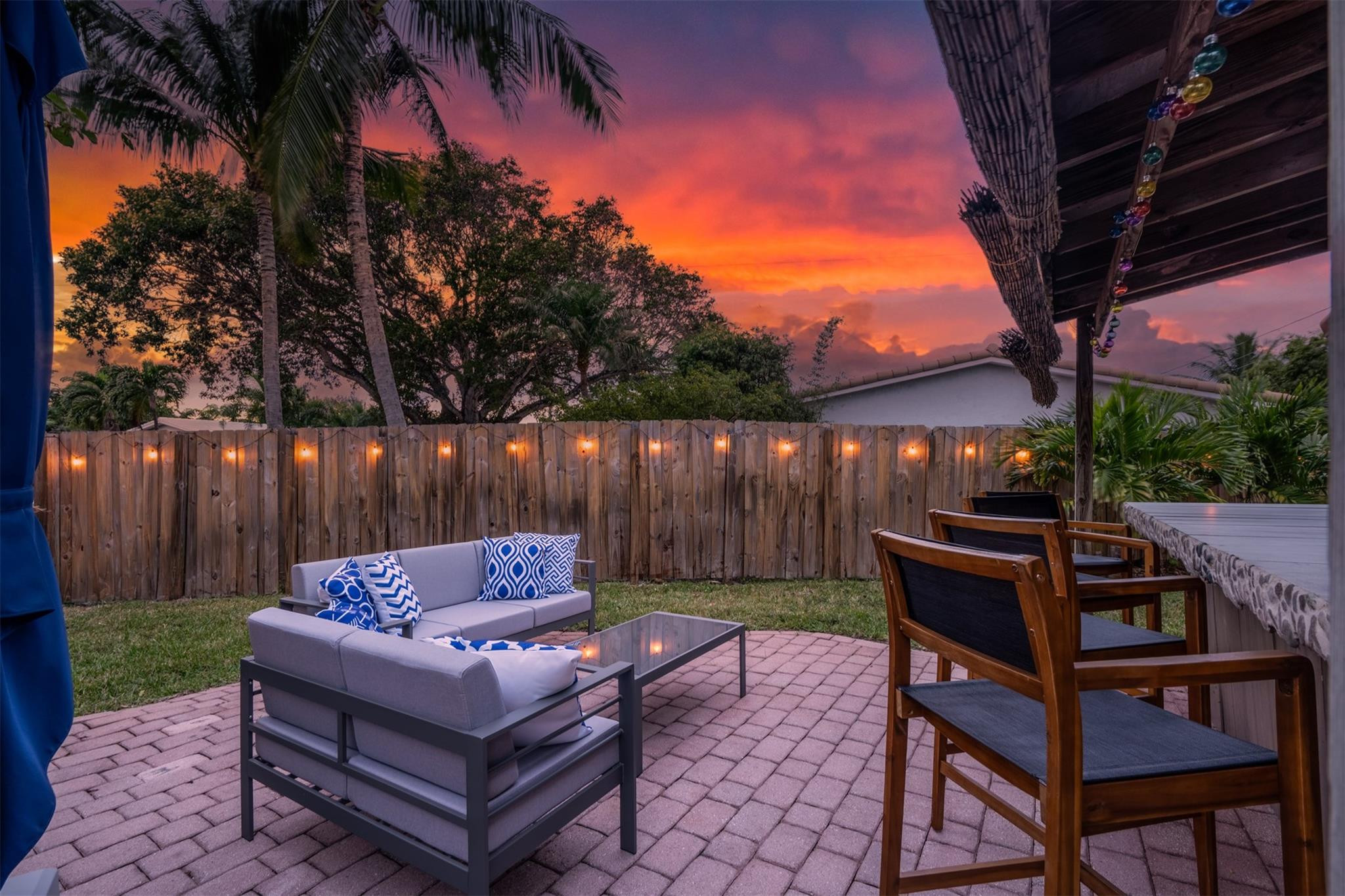 Backyard patio with lounge seating, wooden dining set, string lights, and a vivid sunset sky.