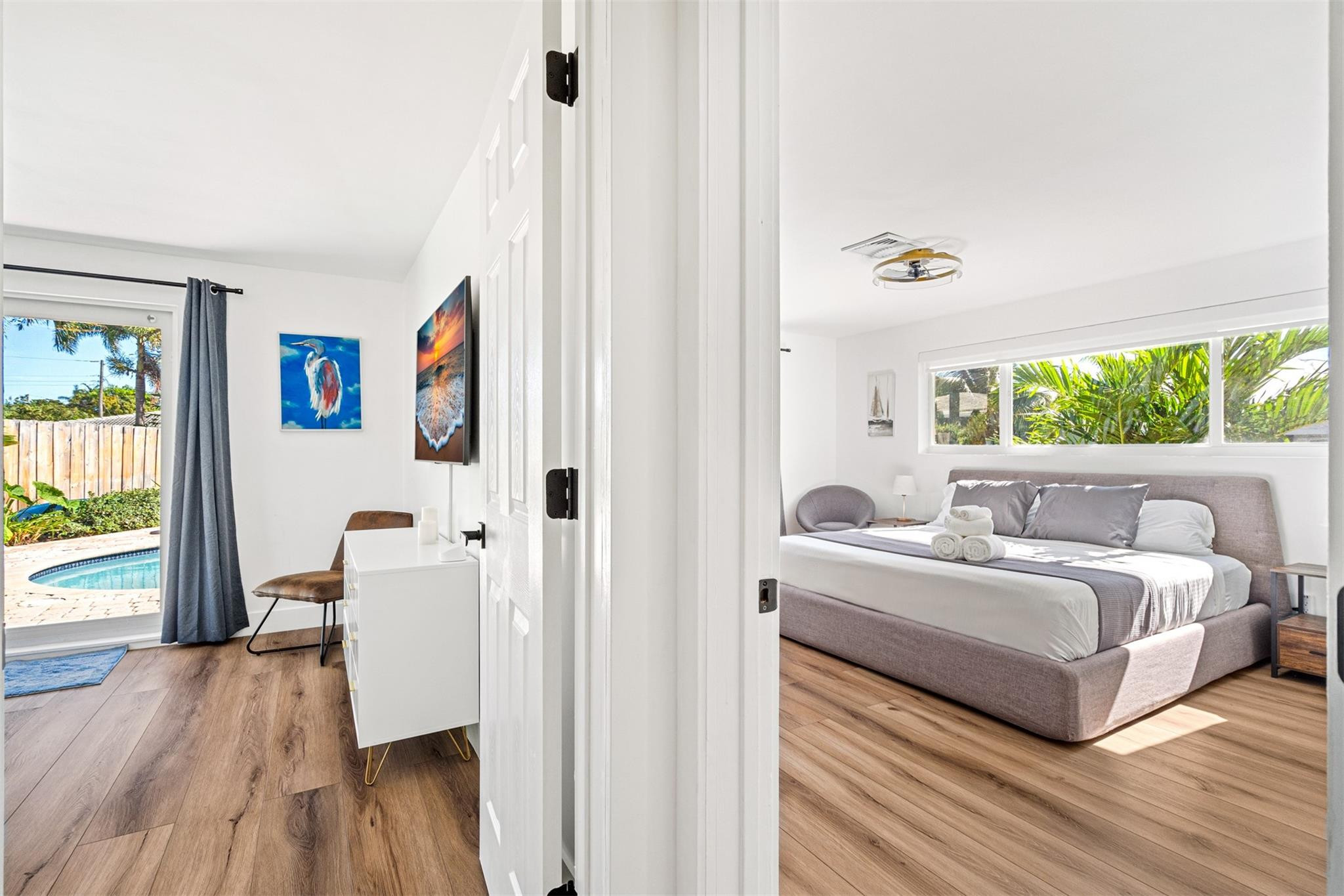 Staged bedroom with a gray upholstered bed, wide window, and tropical greenery outside.