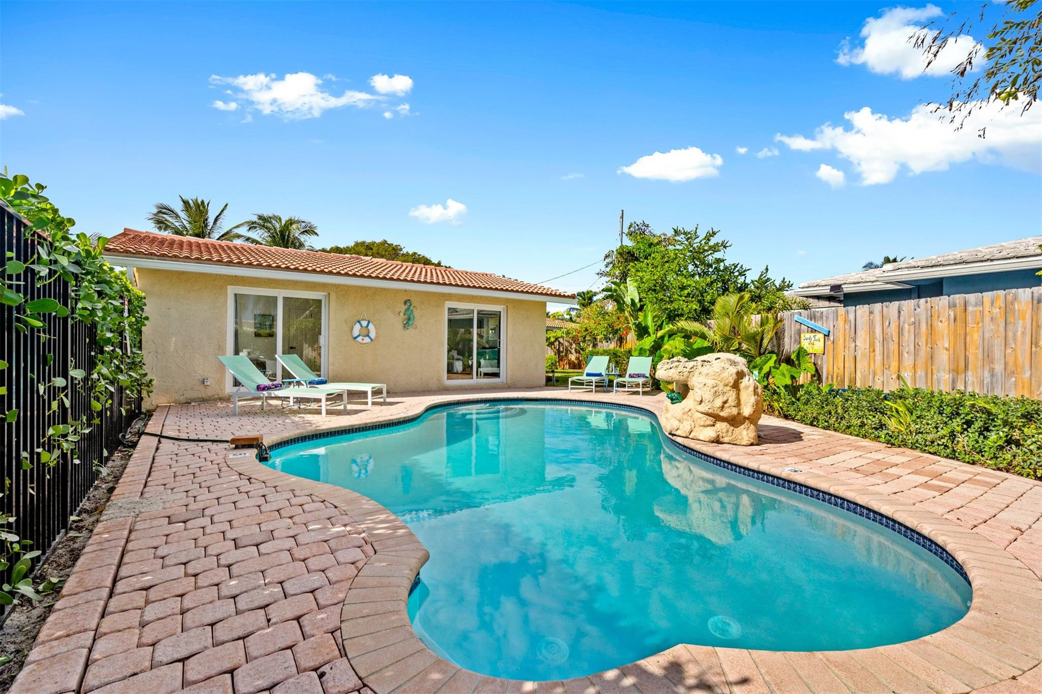 Backyard swimming pool beside a single-story home, with brick pavers, lounge chairs, and tropical landscaping.
