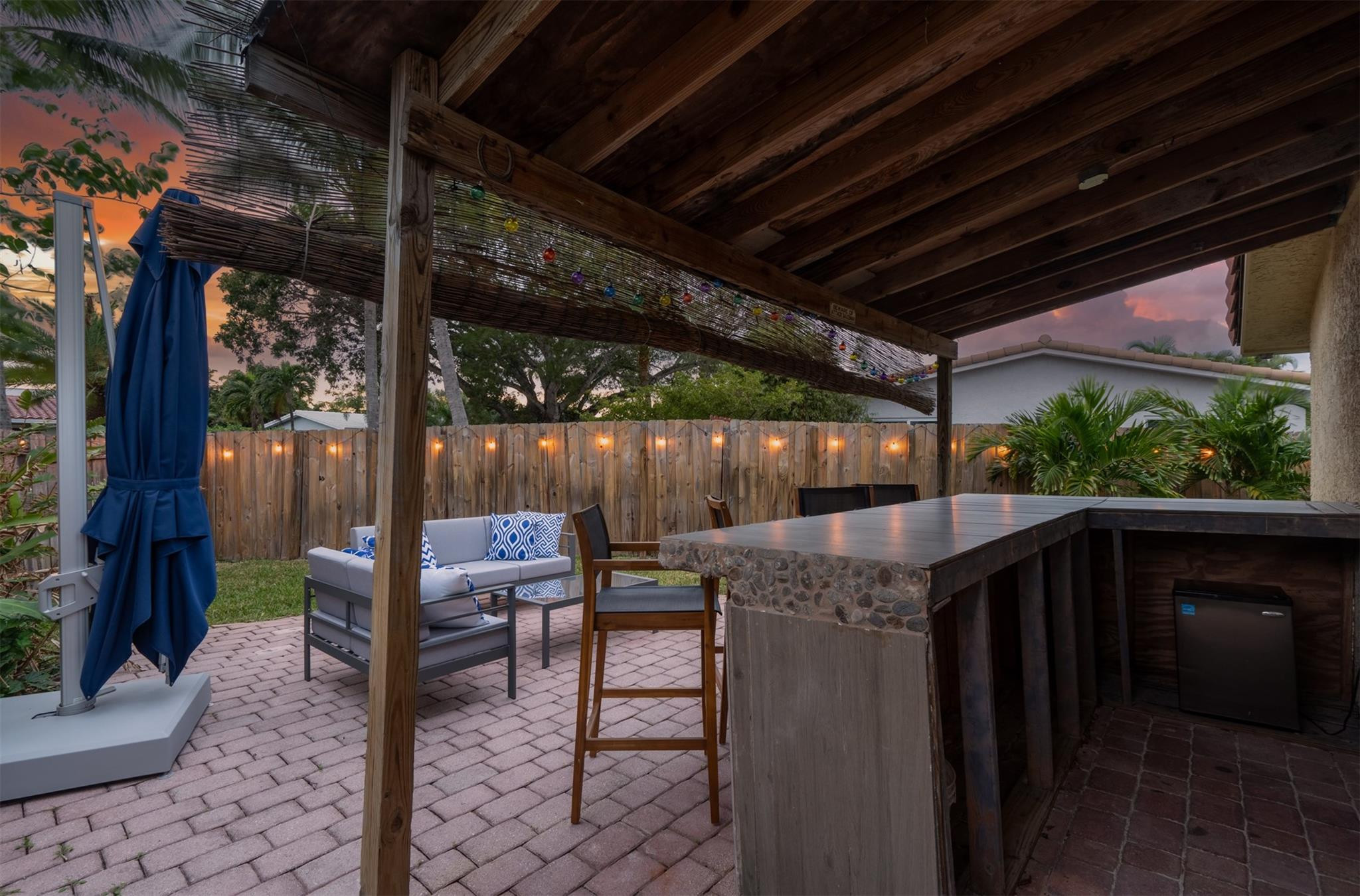 Covered patio with timber pergola, brick pavers, cushioned seating, and built-in bar at dusk.