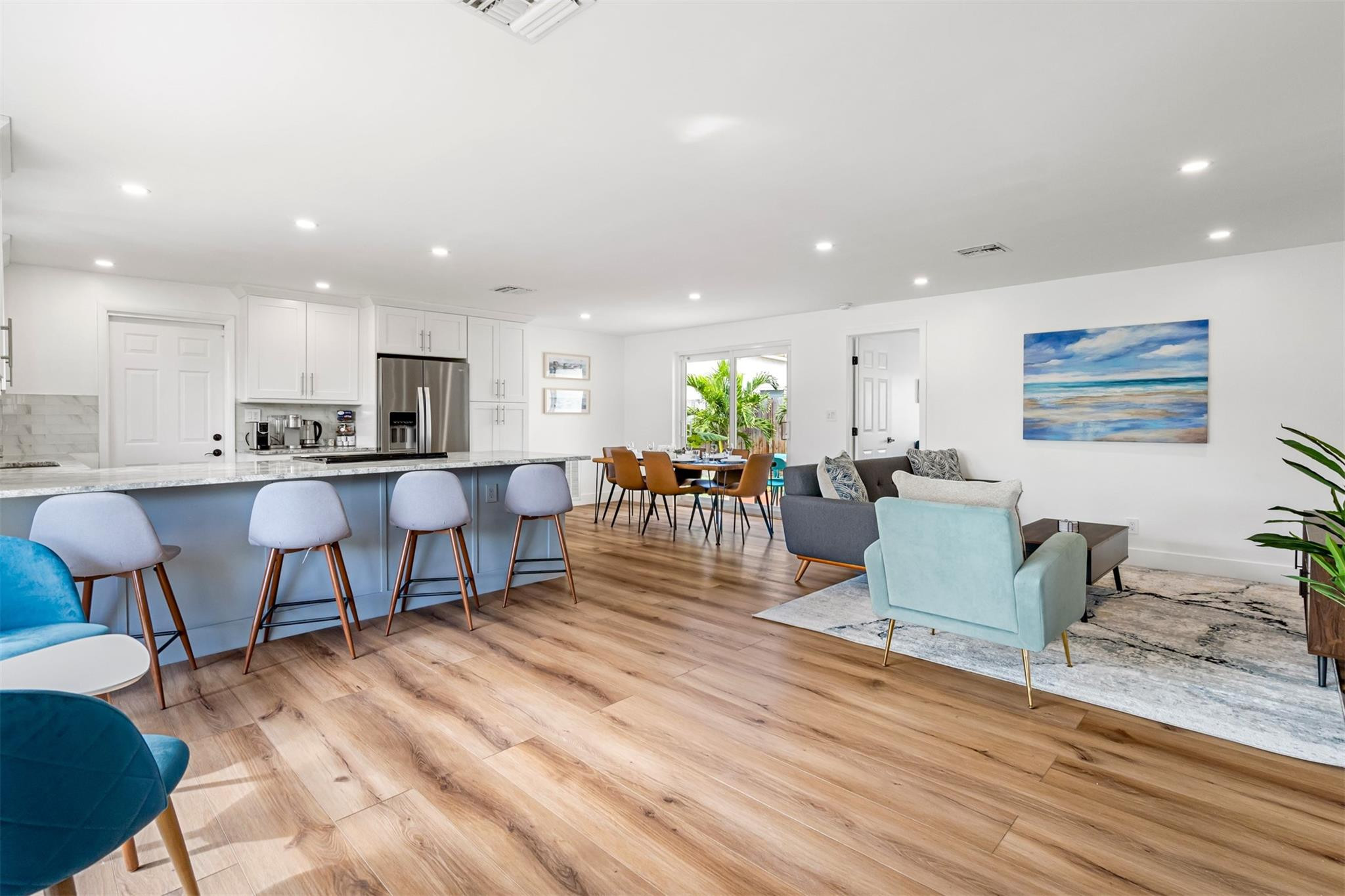Open-plan kitchen and living room with a large island, wood-look floors, and sliding doors to tropical greenery.