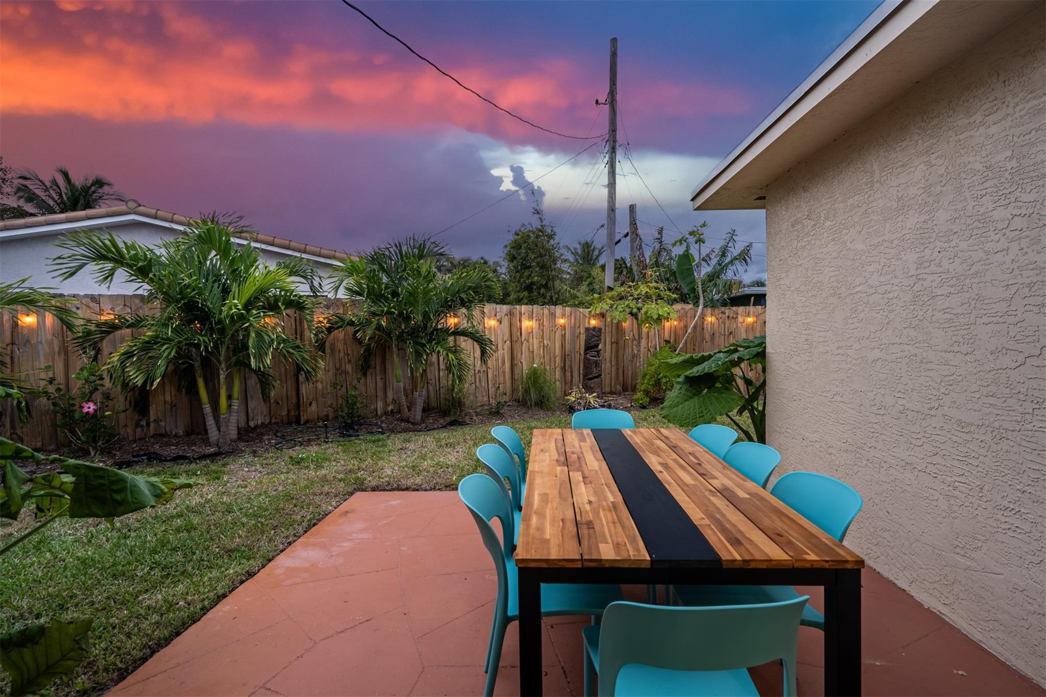 Backyard patio with a wooden dining table, turquoise chairs, palms, and a colorful dusk sky.