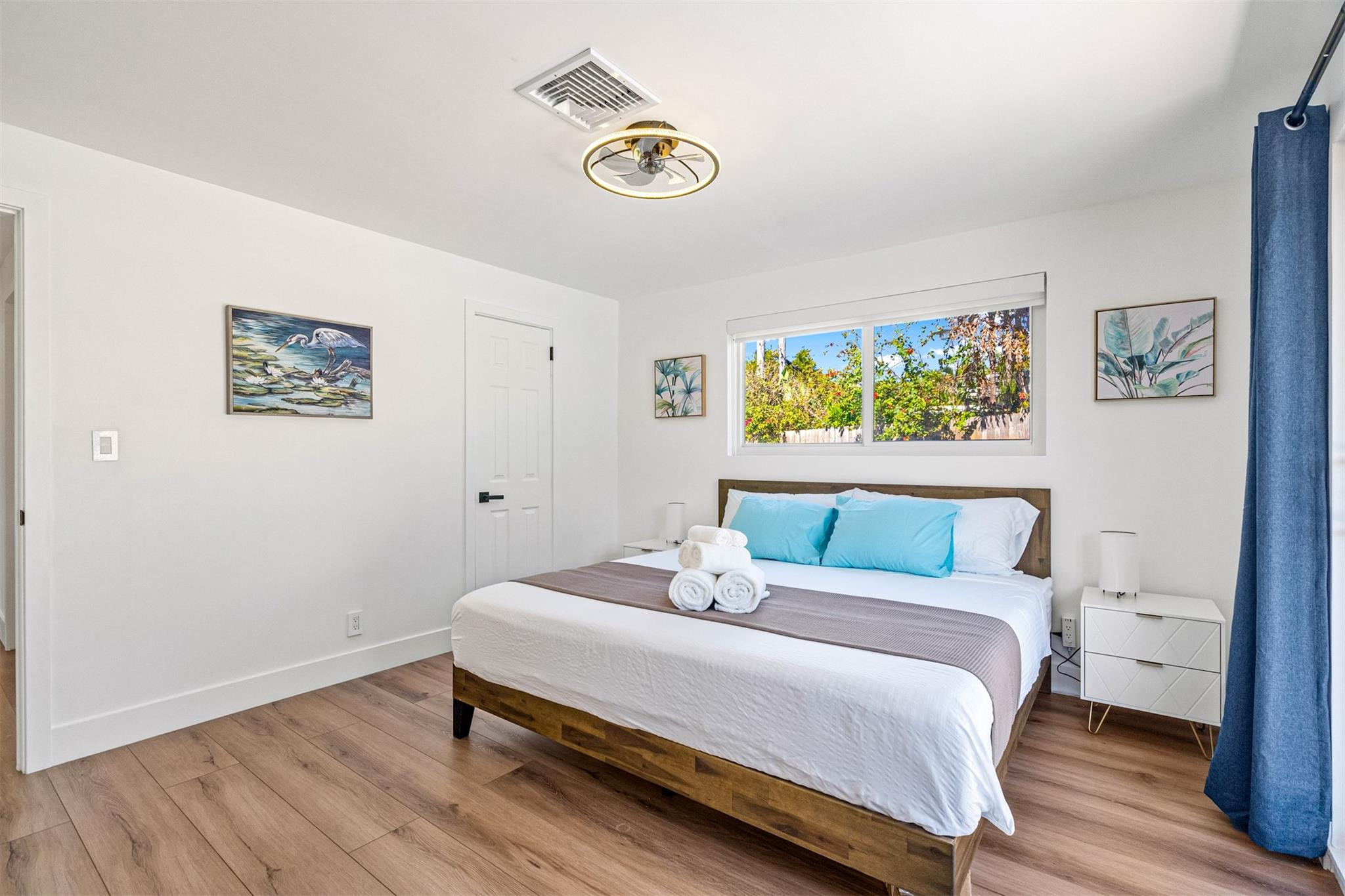 Bright bedroom with a wooden bed, turquoise pillows, rolled towels, and a window overlooking greenery.