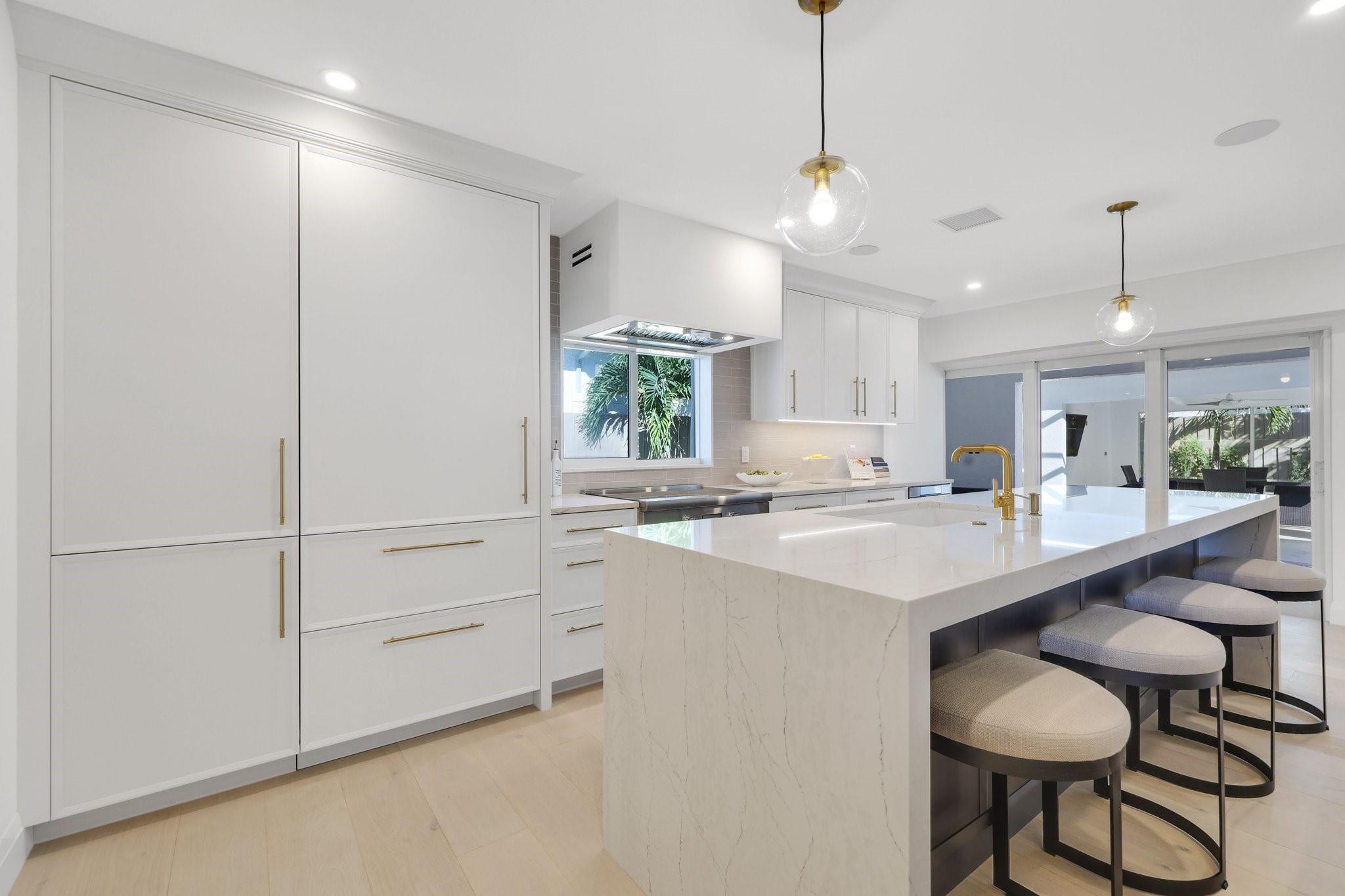 Modern white kitchen with a large stone island and stools beside sliding glass doors to an outdoor dining area.