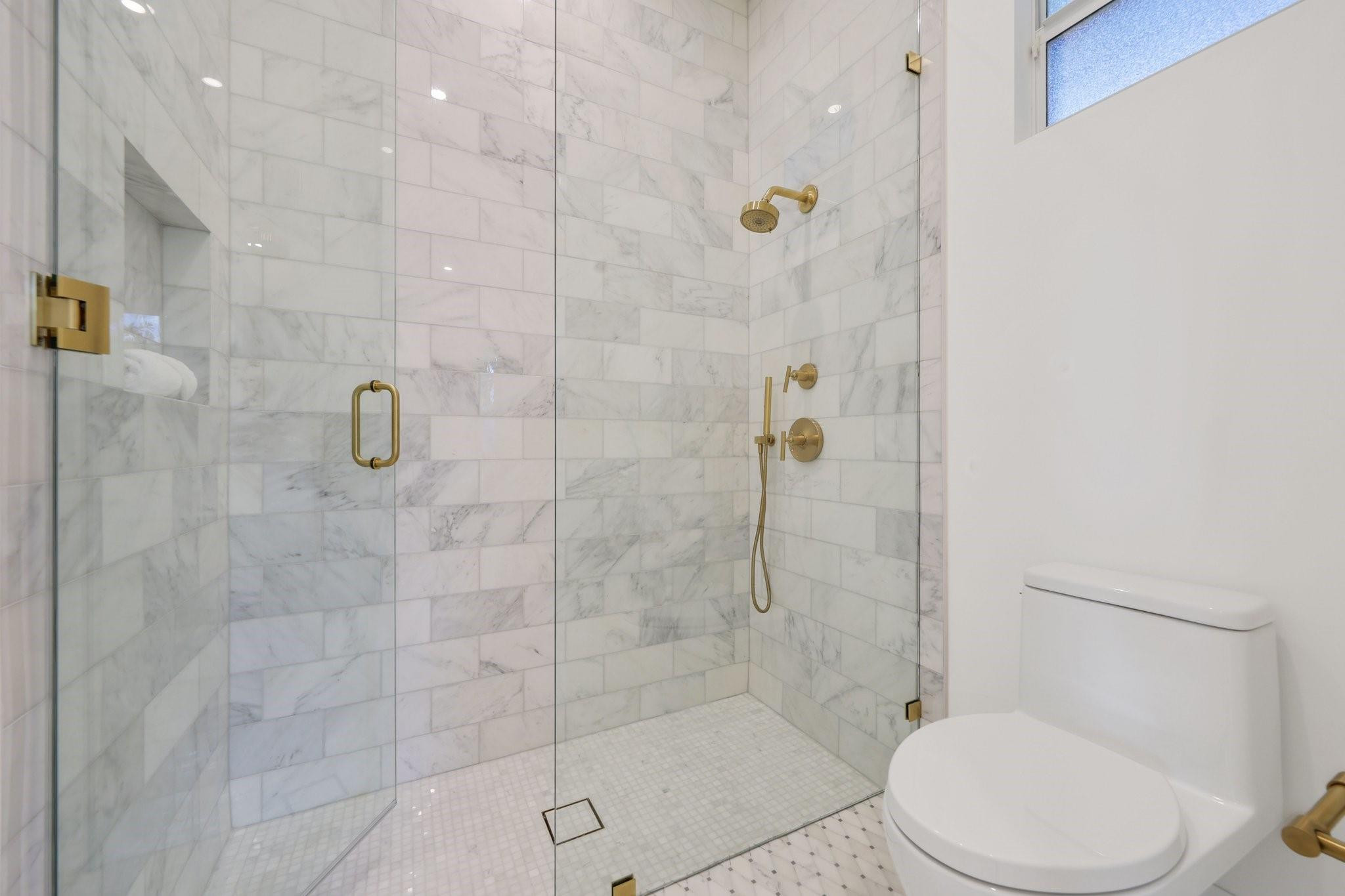 Bathroom with a glass-enclosed marble-tile shower and brass fixtures beside a toilet.