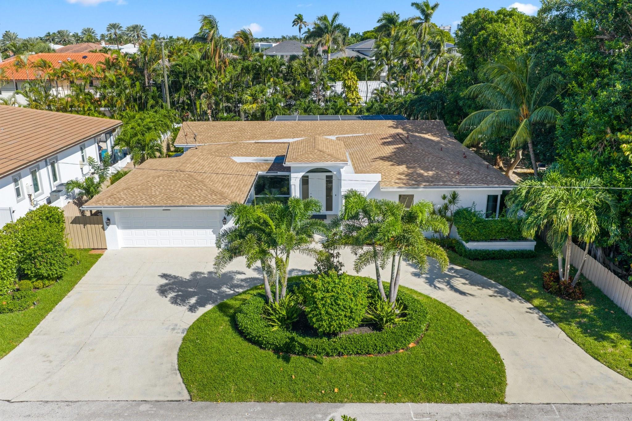 Aerial view of a white house with a two-car garage, curved driveway, and palm-lined landscaping.