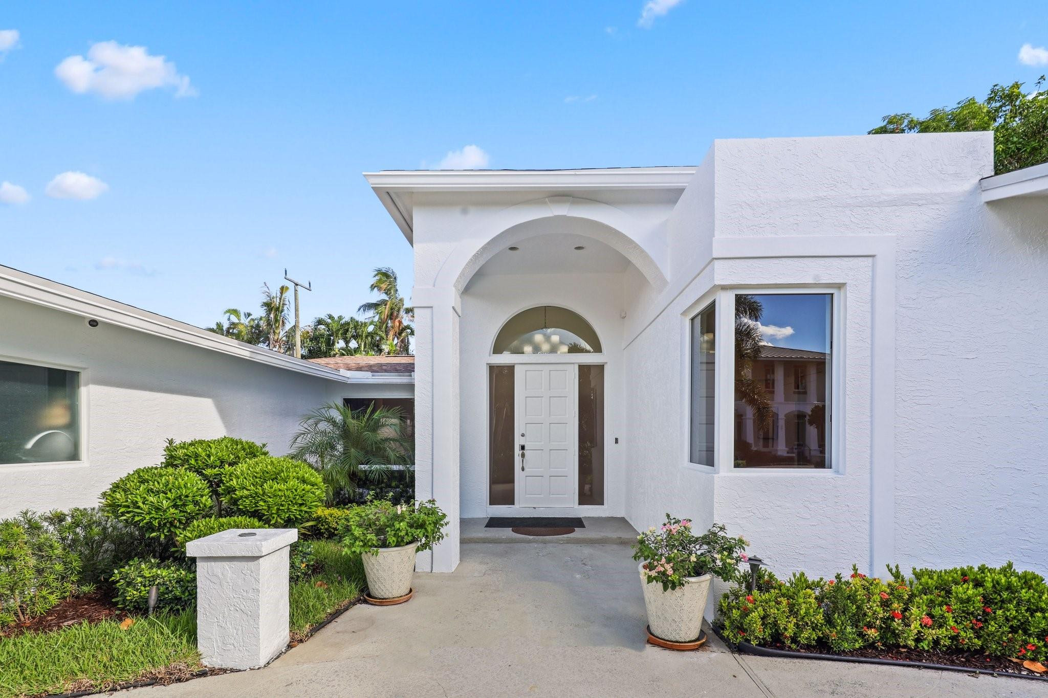 White stucco home with an arched front entry, potted plants, and landscaped shrubs.
