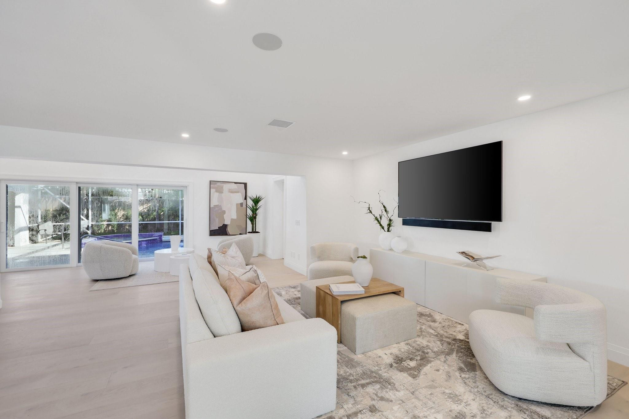 Staged living room with white seating, a wall-mounted television, and sliding doors overlooking a pool.