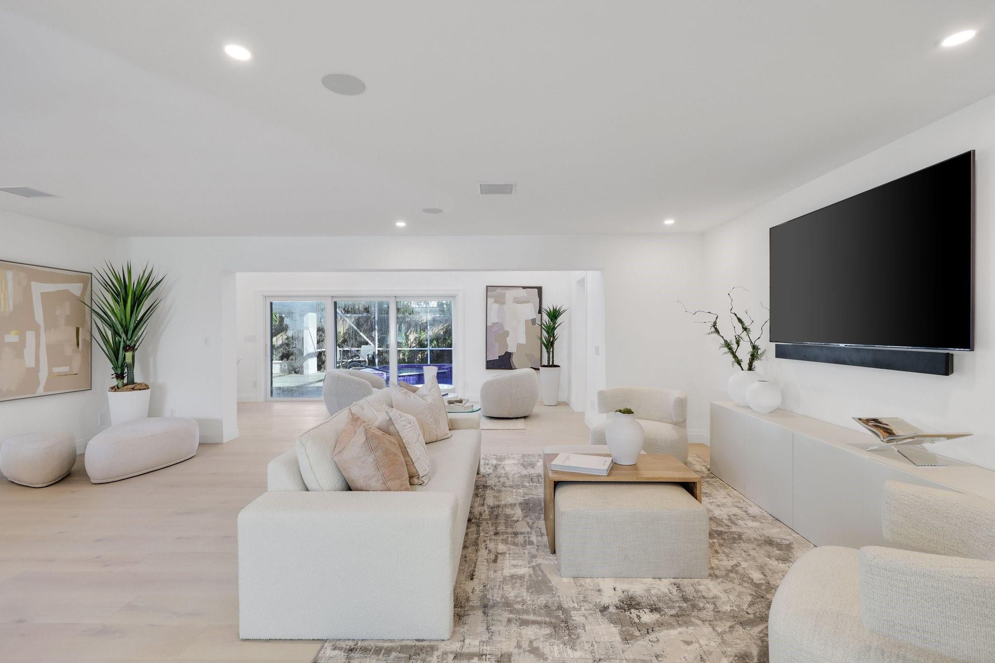 Bright living room with white seating, a large wall-mounted TV, and sliding doors overlooking a pool.