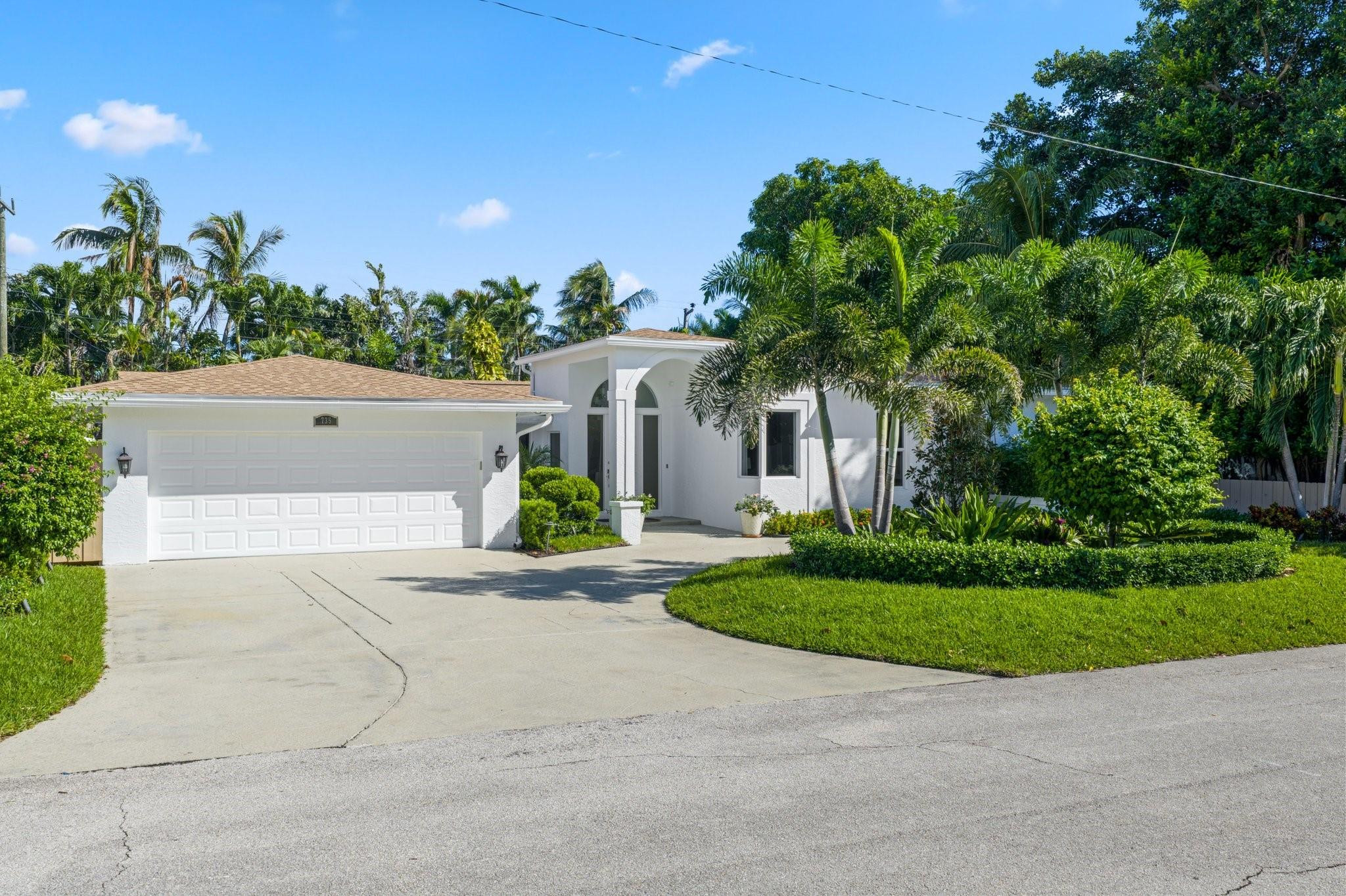 White home with a two-car garage, arched entry, curved driveway, and tropical landscaping.