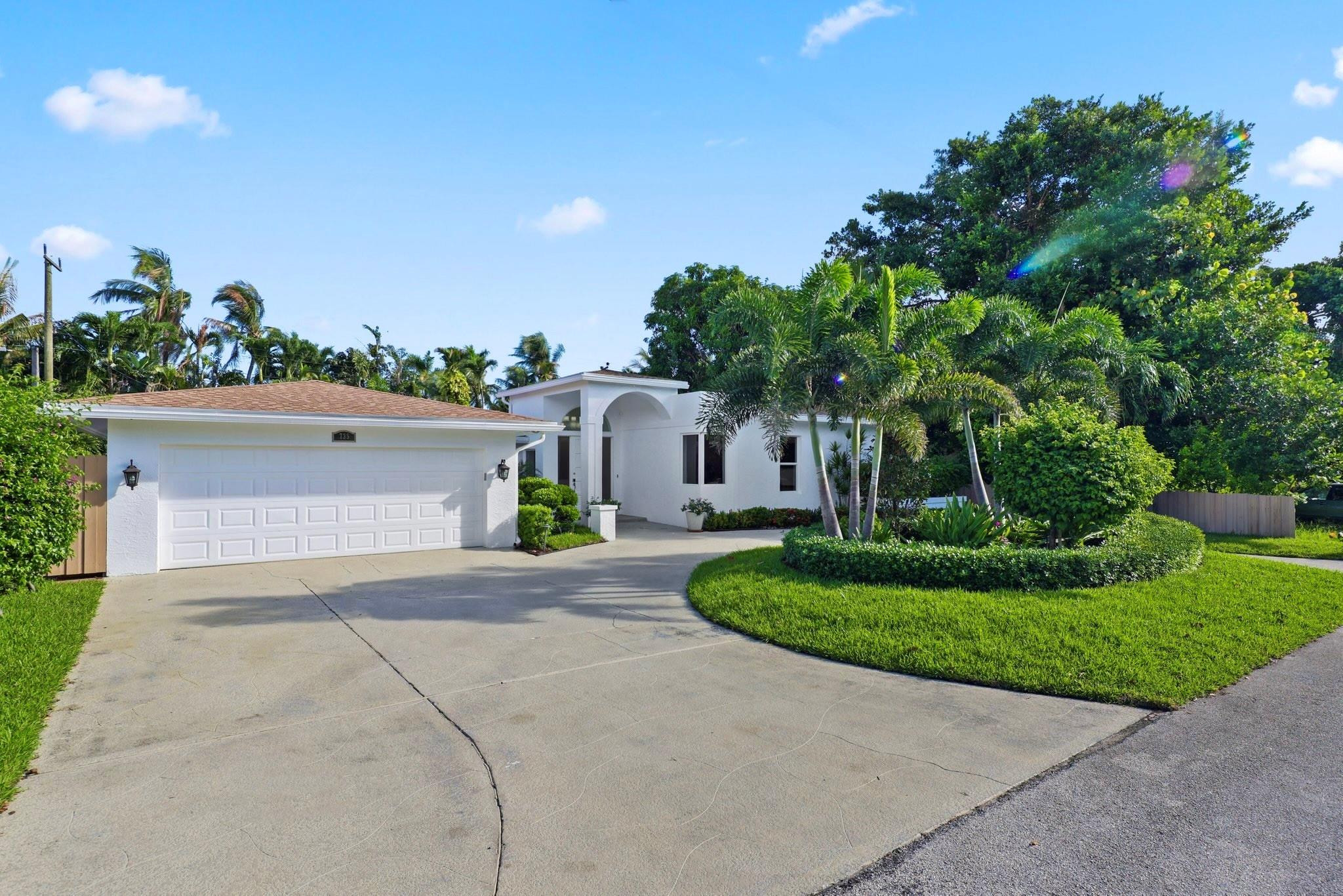 White home with an arched entry, two-car garage, and tropical landscaping.