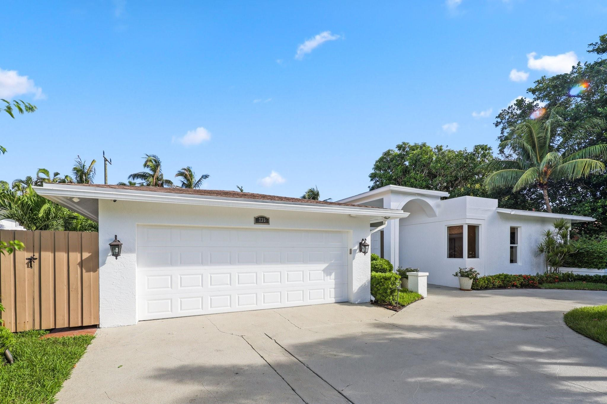 White stucco home with a wide two-car garage, curved entry detail, and tropical landscaping.