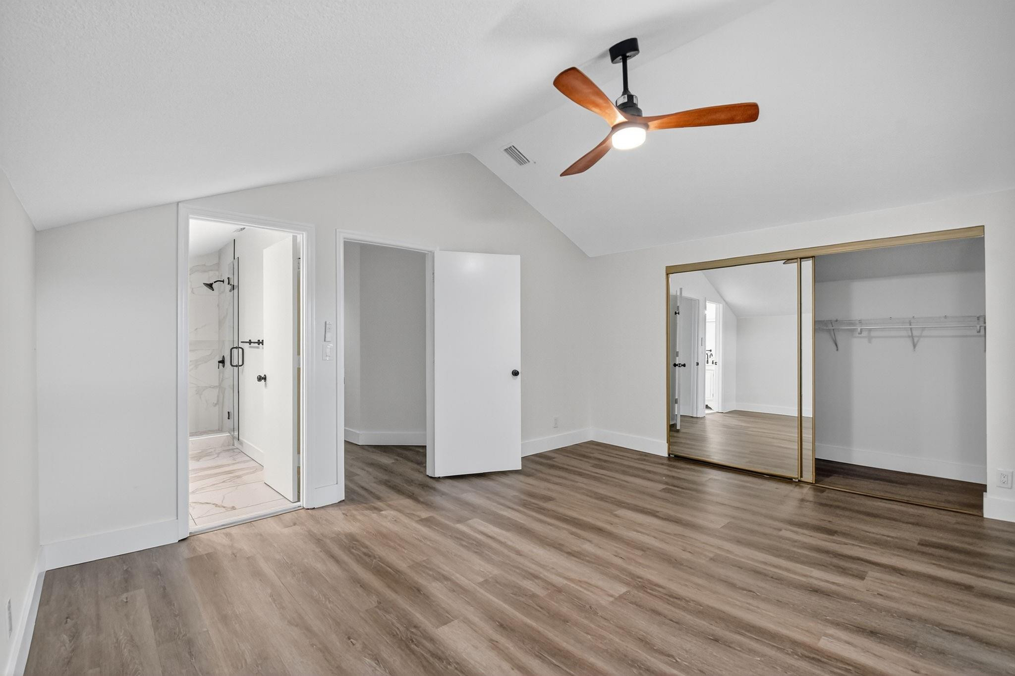 Empty bedroom with vaulted ceiling, wood-blade fan, mirrored closet doors, and an adjoining bathroom.
