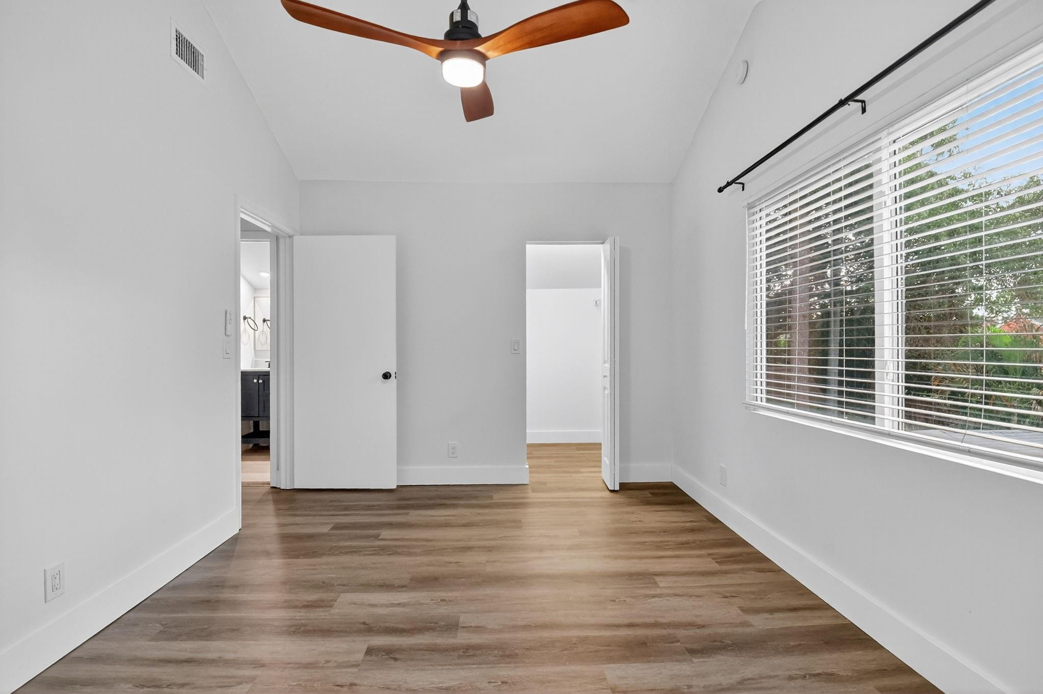 Empty bedroom with plank flooring, a wood-blade ceiling fan, and a large window overlooking greenery.