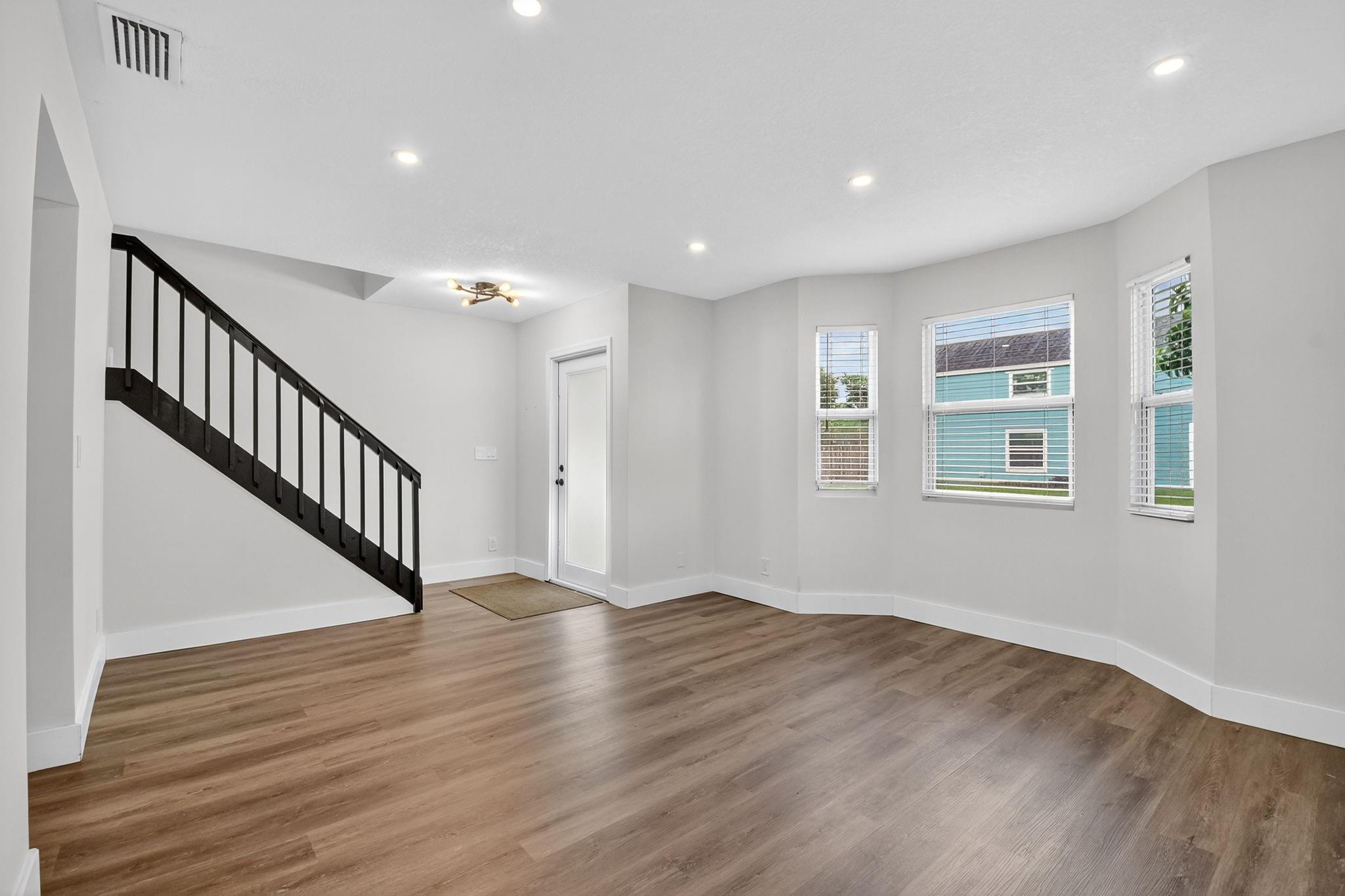 Empty living area with wood-look floors, a black-railed staircase, and three windows with blinds.