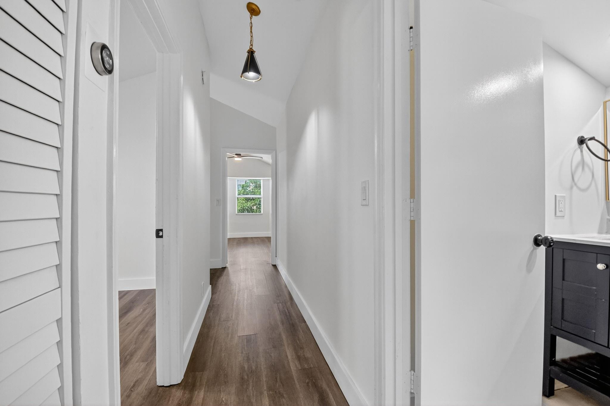 Hallway with vaulted white ceiling, wood-look floors, black pendant light, and a bathroom vanity at right.