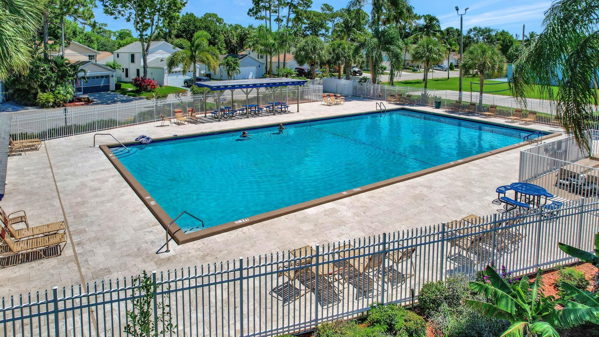 Outdoor community pool with a fenced paved deck, shaded picnic tables, and palm trees.