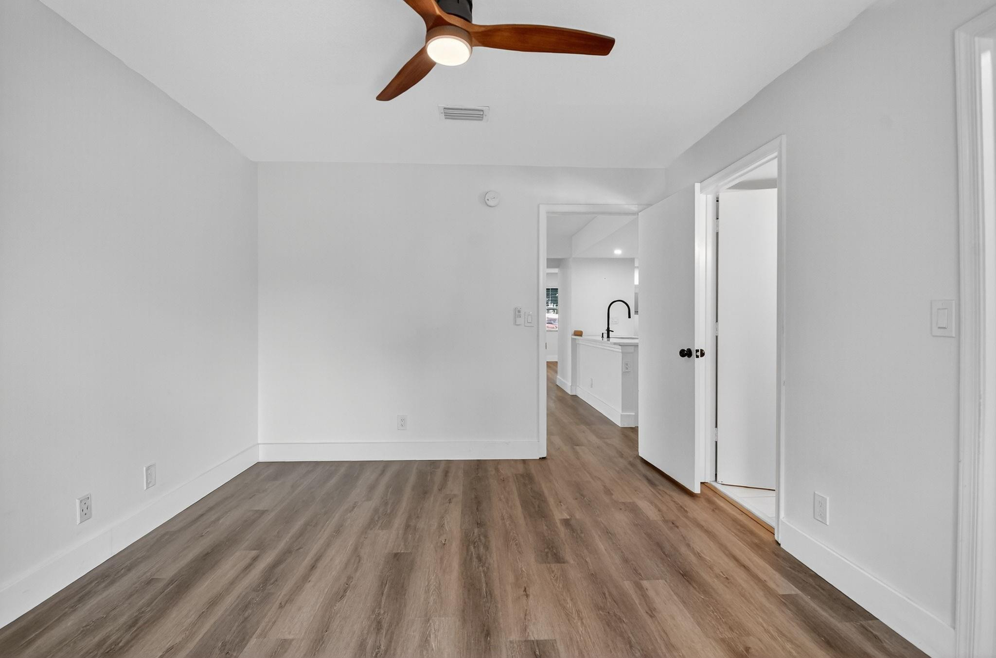 Unfurnished room with wood-look floors and ceiling fan, opening toward a bright kitchen.