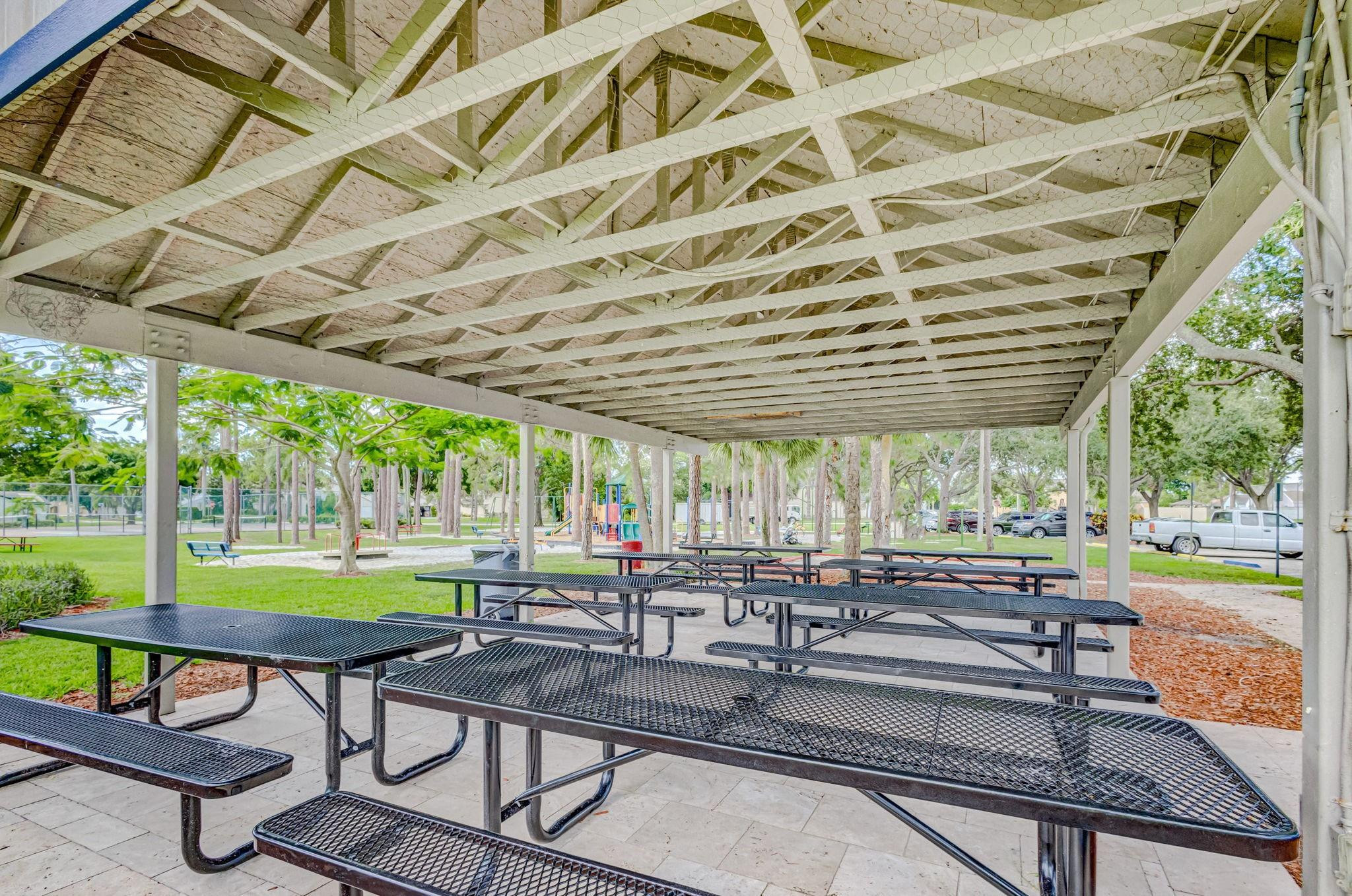 Covered picnic pavilion with black metal tables overlooking a leafy park and playground.
