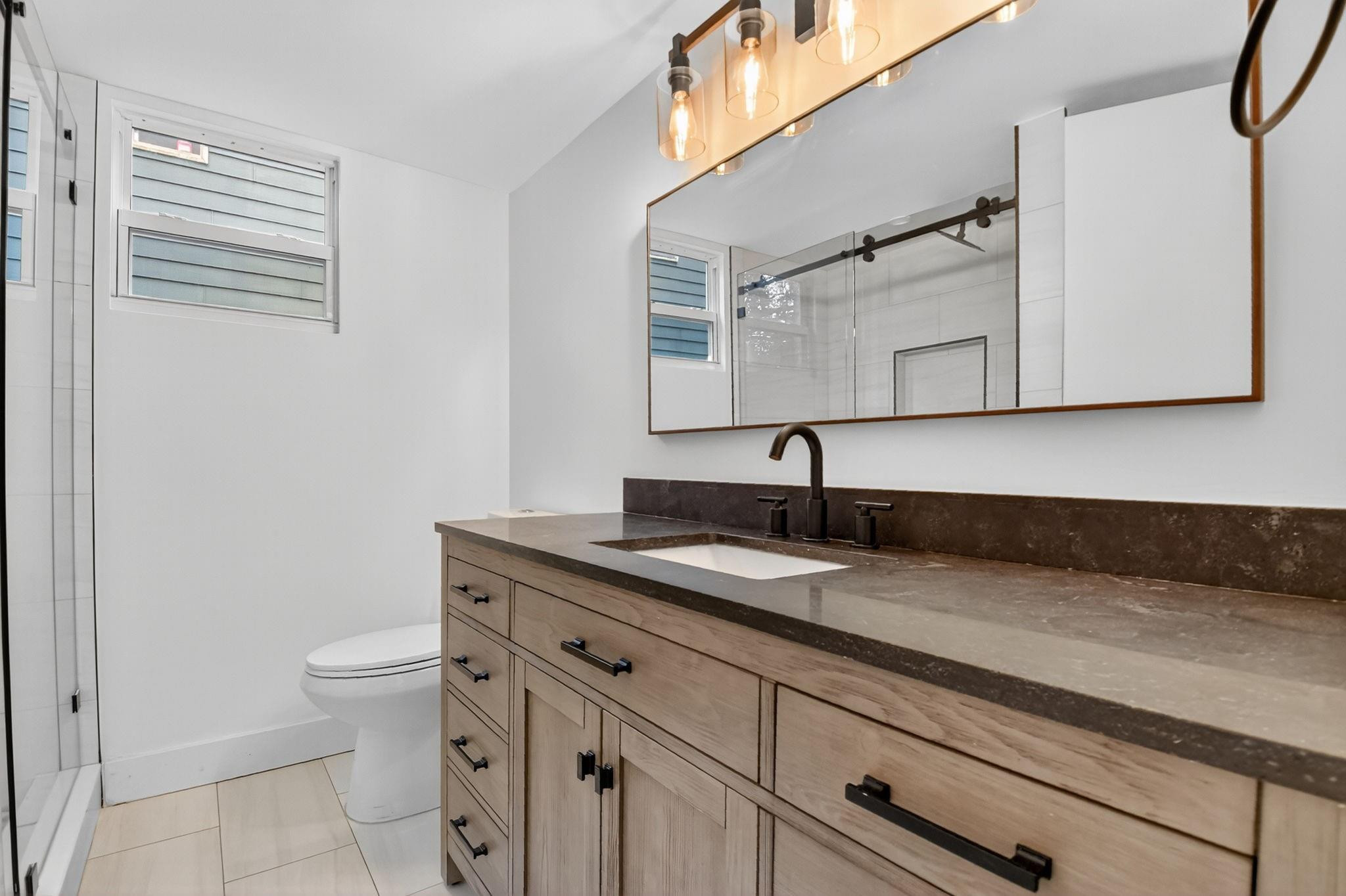 Modern bathroom with a long wood vanity, dark stone counter, glass shower, and black fixtures.
