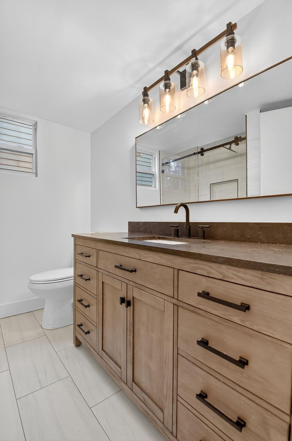Bathroom vanity with light wood cabinetry, dark countertop, wide mirror, and bronze lighting.