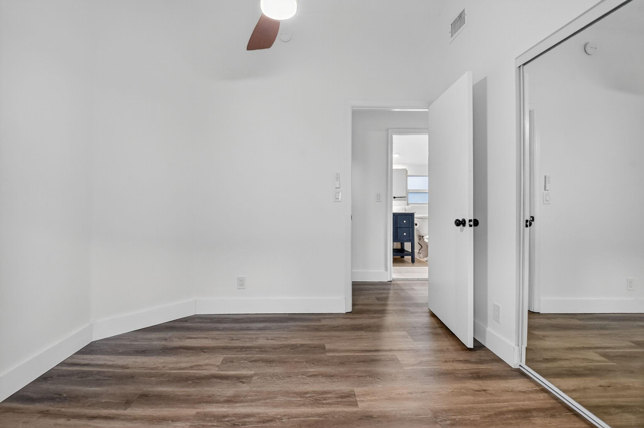 Vacant bedroom with wood-look flooring, mirrored closet doors, and an open doorway to a bathroom.