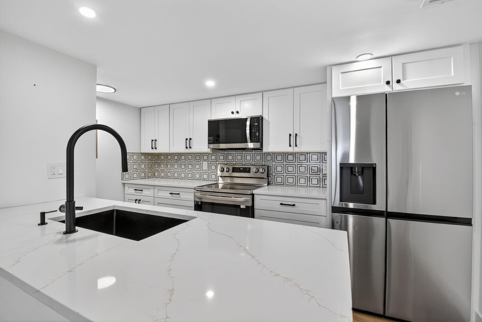 Renovated kitchen with white cabinets, patterned tile backsplash, stainless appliances, and a large island sink.