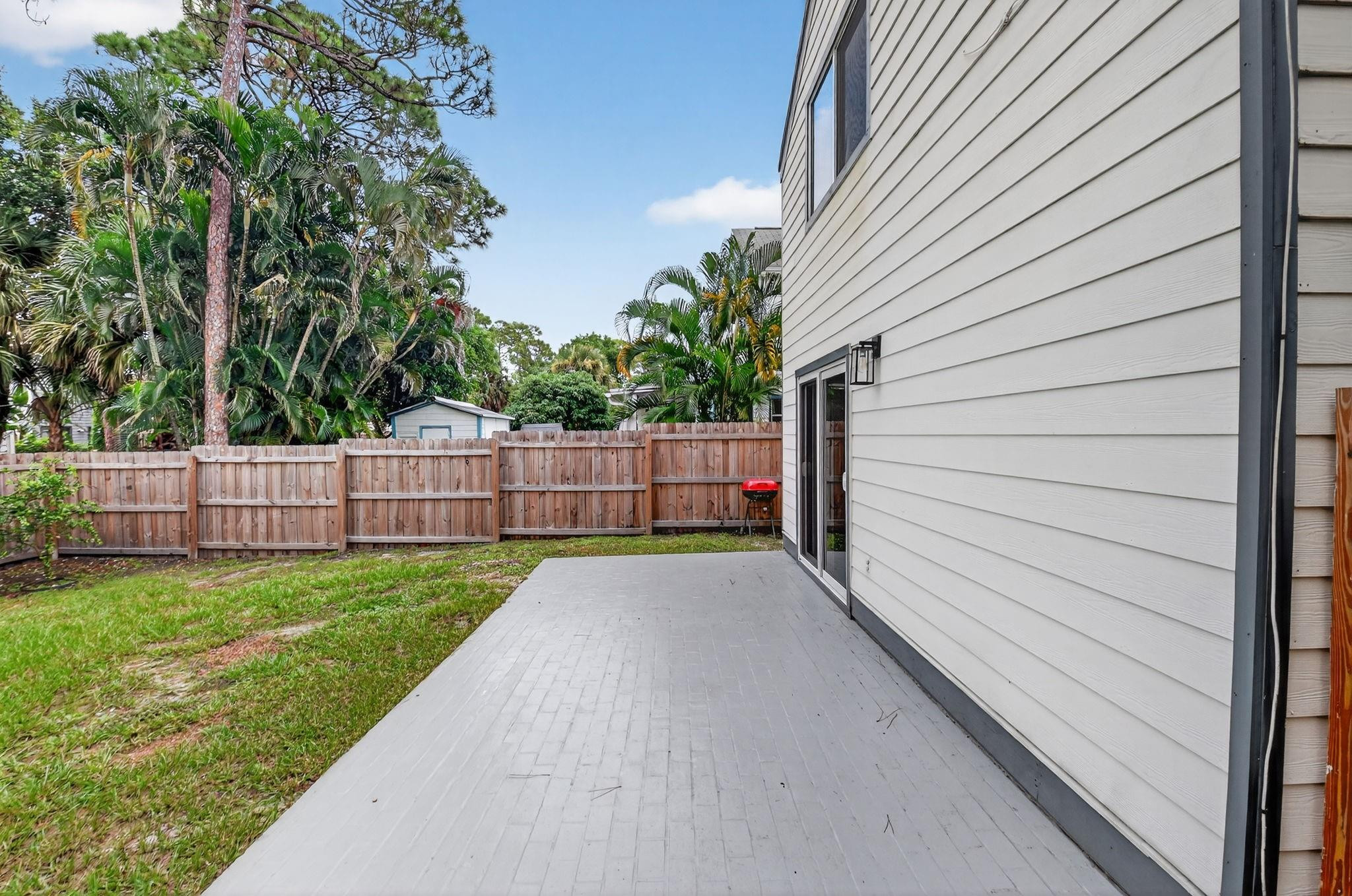 Paved backyard patio beside the home, with a fenced lawn and small red grill.