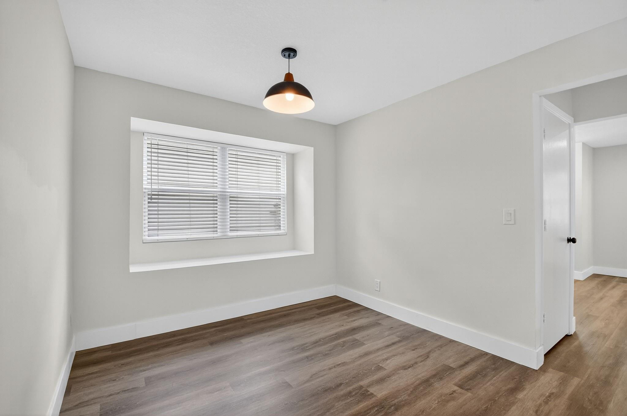 Empty room with wood-look flooring, a recessed window with blinds, and a black pendant light.