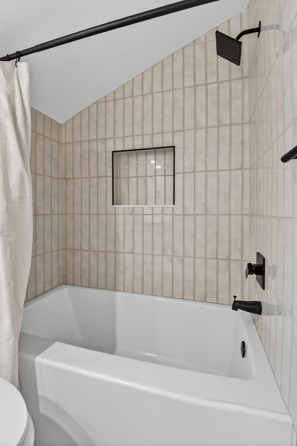 Bathroom bathtub with beige tile surround, black fixtures, and recessed wall niche.