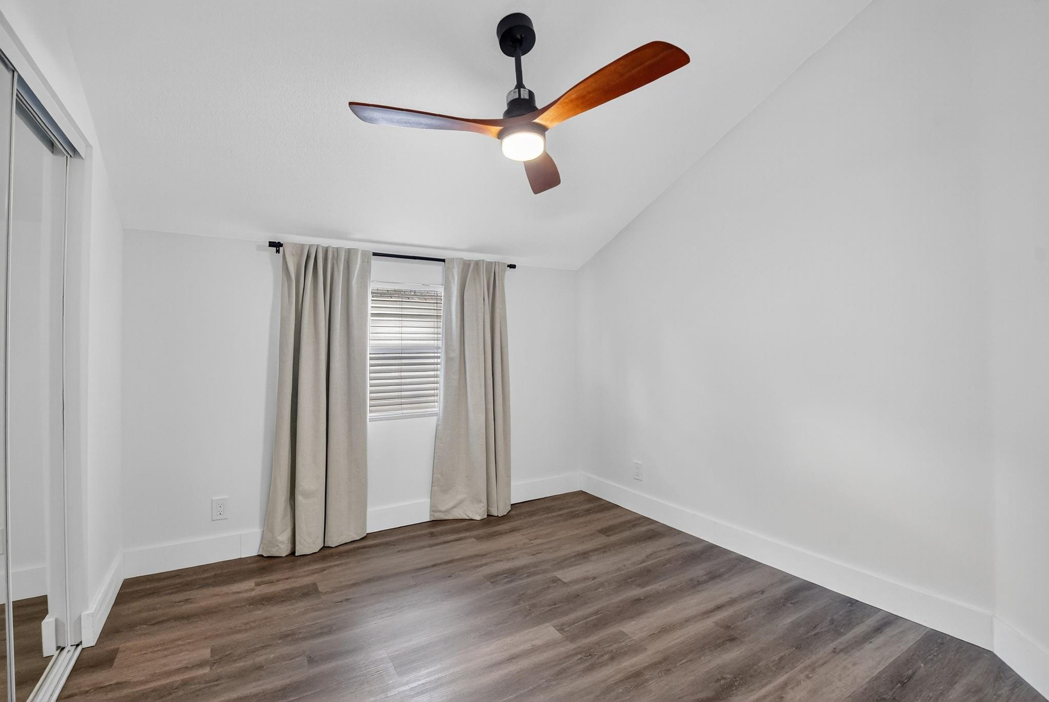 Empty bedroom with wood-look flooring, curtained window, mirrored closet doors, and a wood-blade ceiling fan.