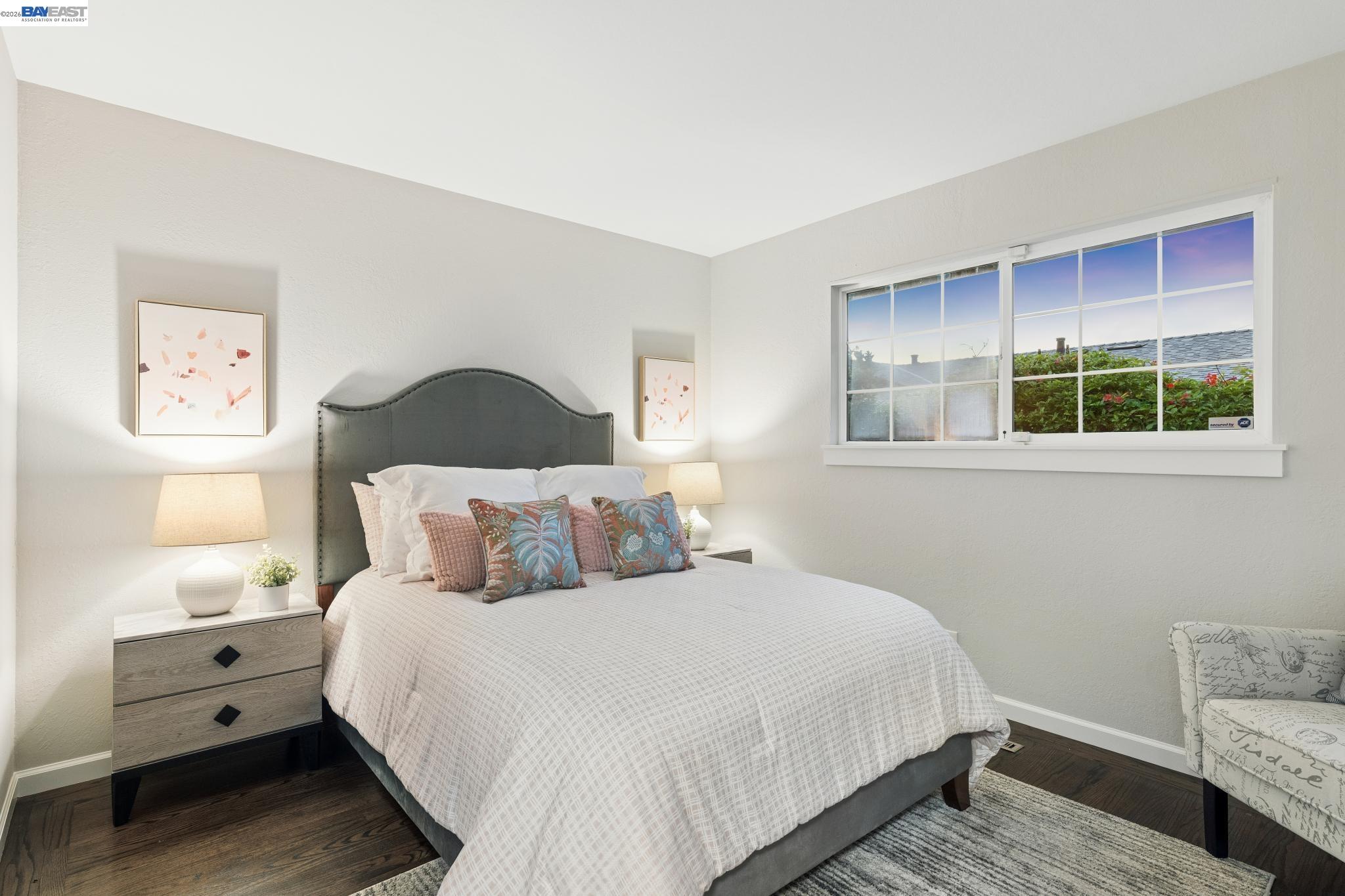 Furnished bedroom with a gray upholstered bed, colorful pillows, and a large window overlooking shrubs.