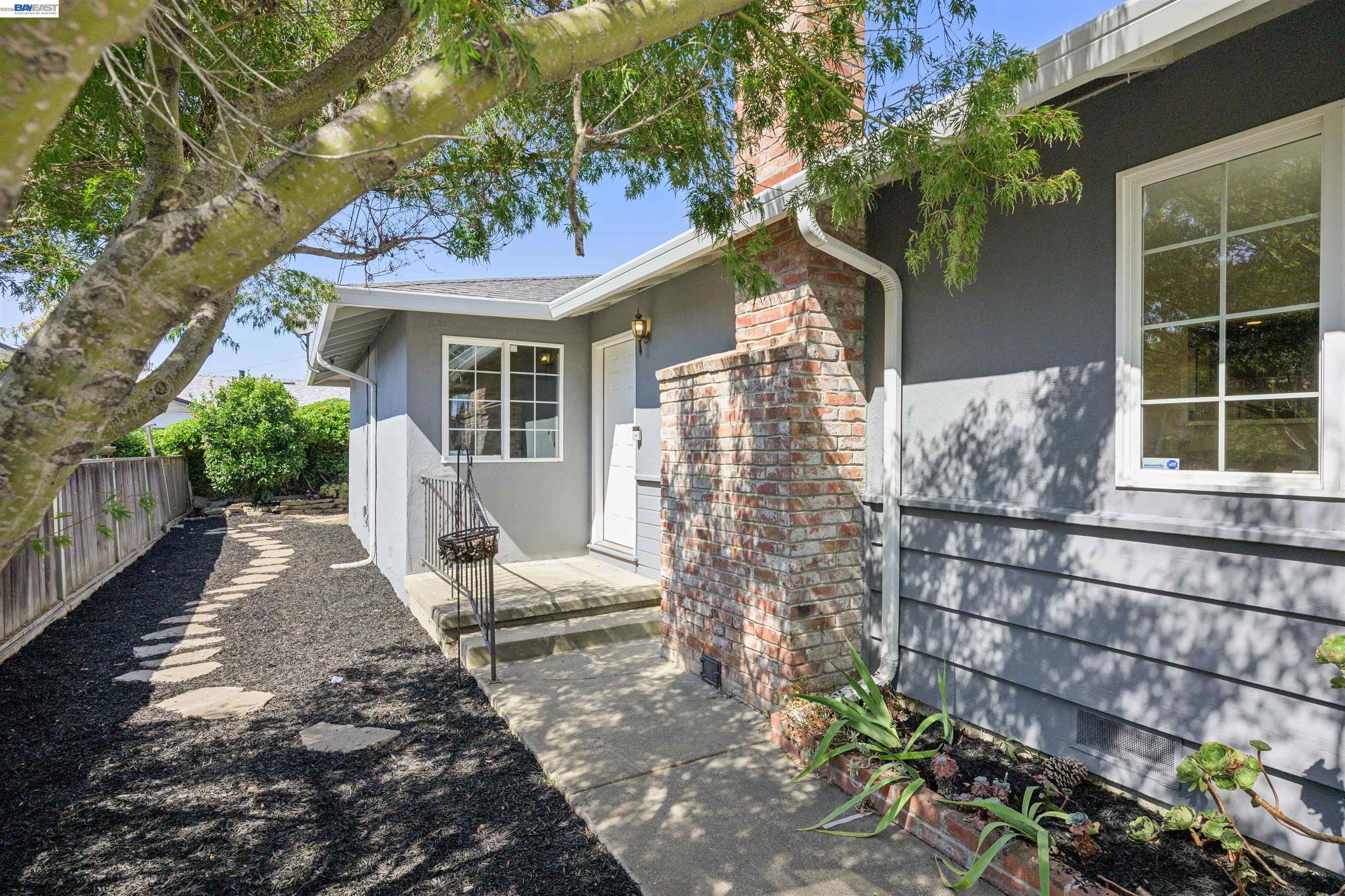 Gray house entrance with a brick column, covered porch, and stepping-stone path beneath a leafy tree.