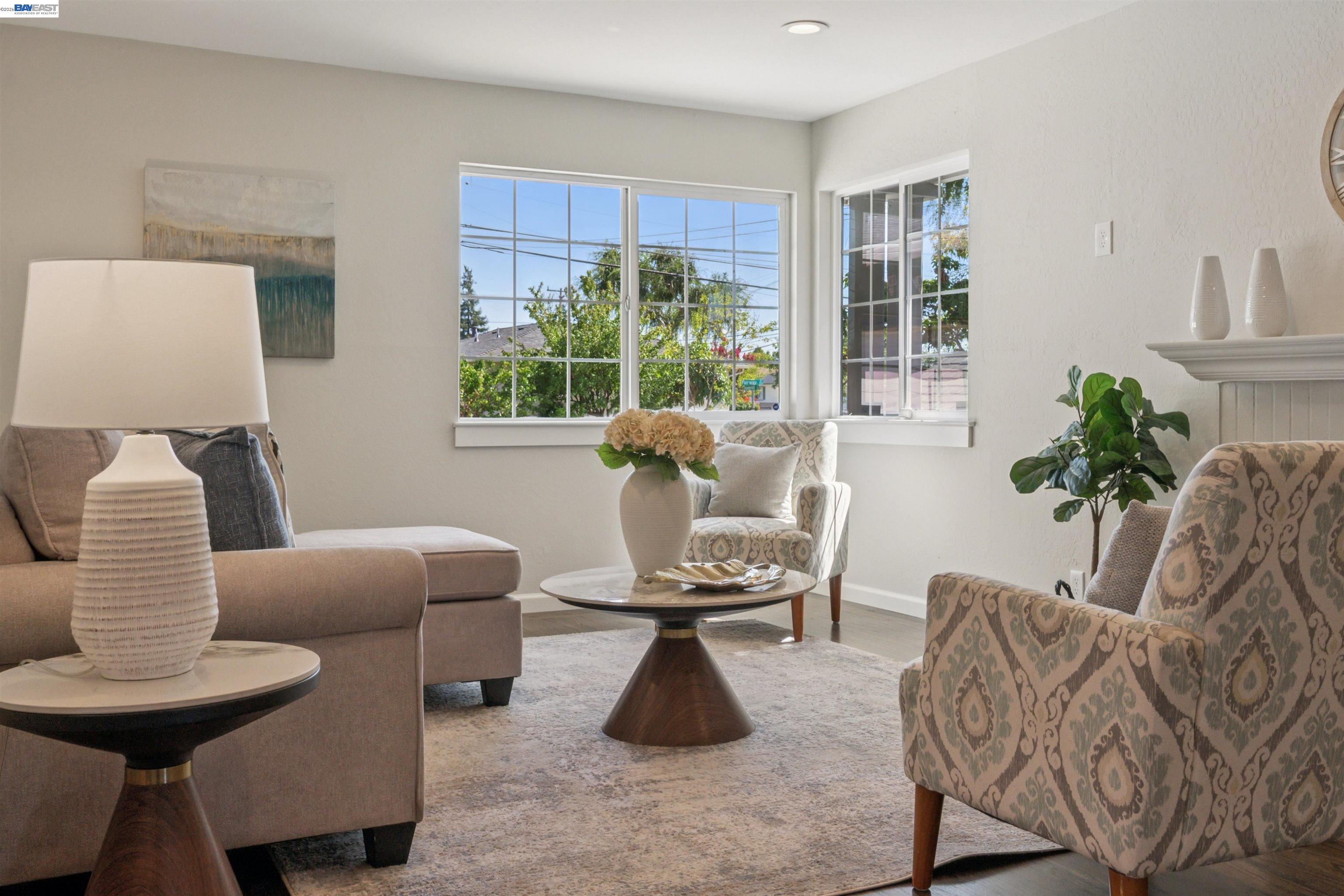 Staged living room with large bright windows, a taupe sofa, patterned armchairs, and a fireplace.