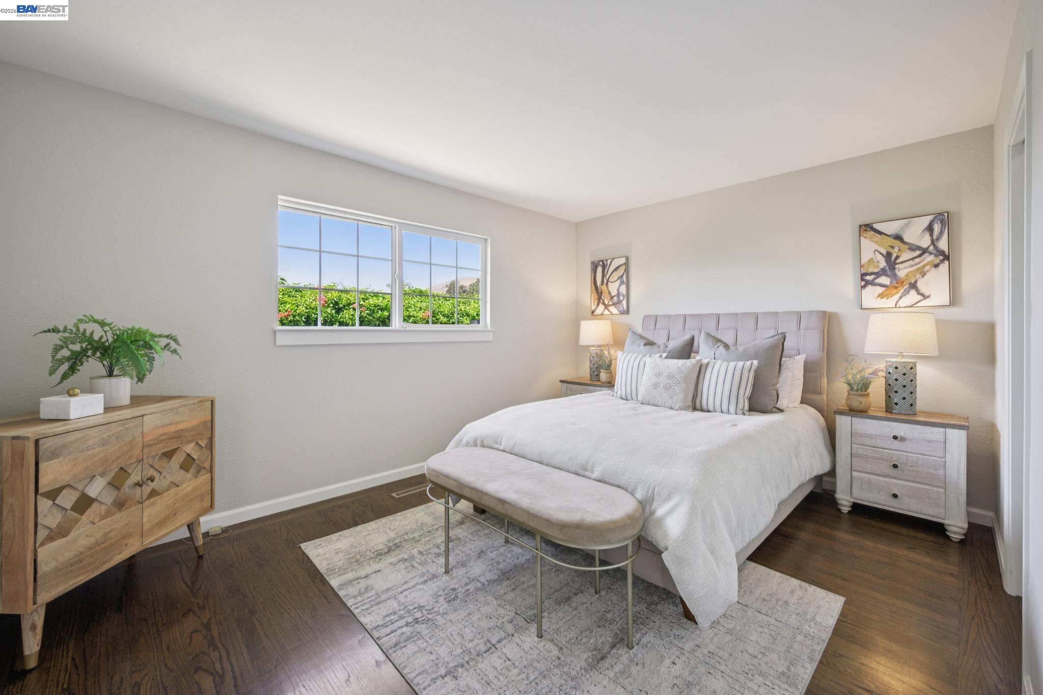 Staged bedroom with a large bed, window overlooking greenery, and dark wood floors.