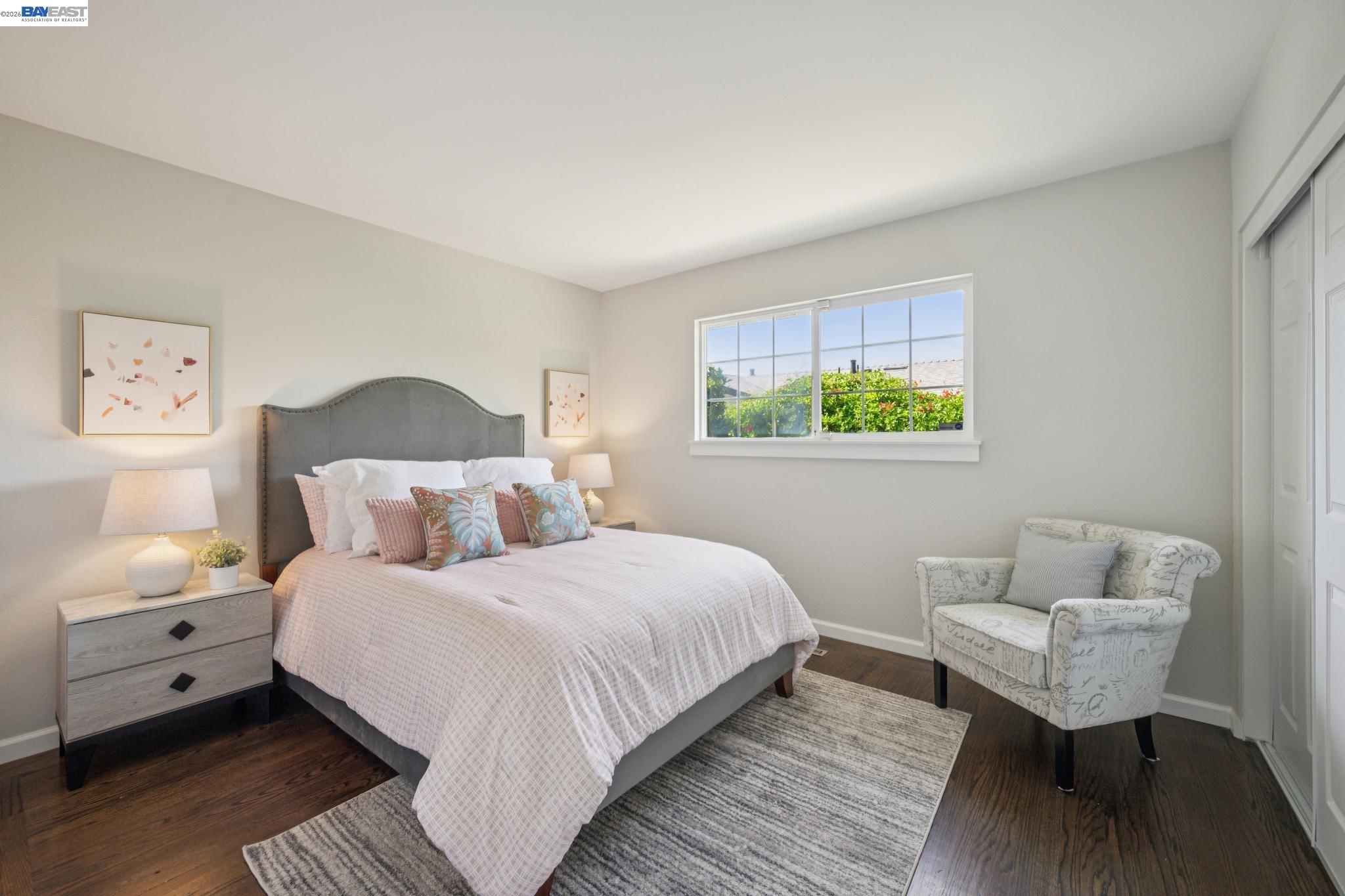 Staged bedroom with a gray upholstered bed, patterned armchair, bright window, and dark wood flooring.