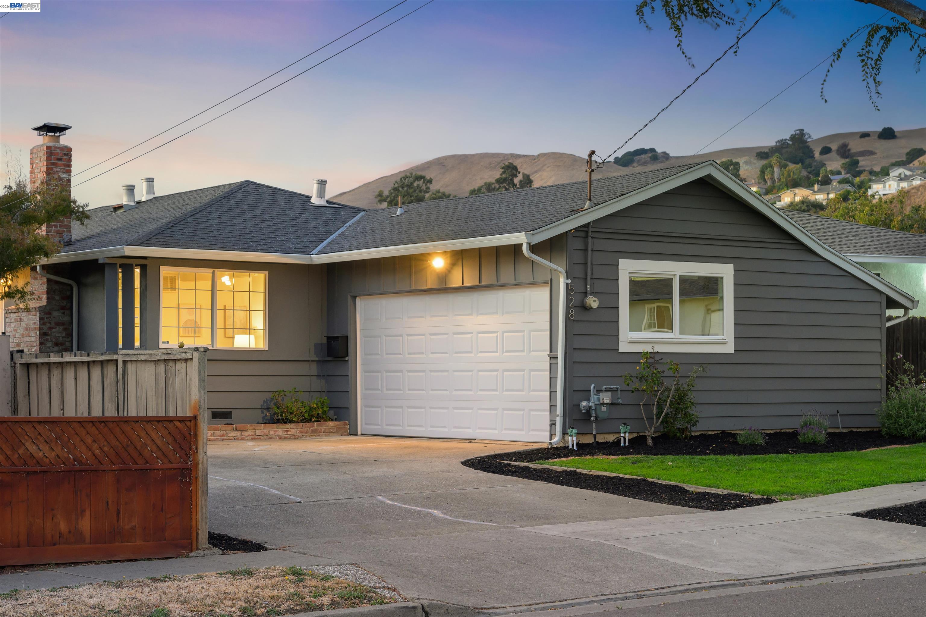 Gray single-story home with a white garage door and warmly lit windows at dusk.