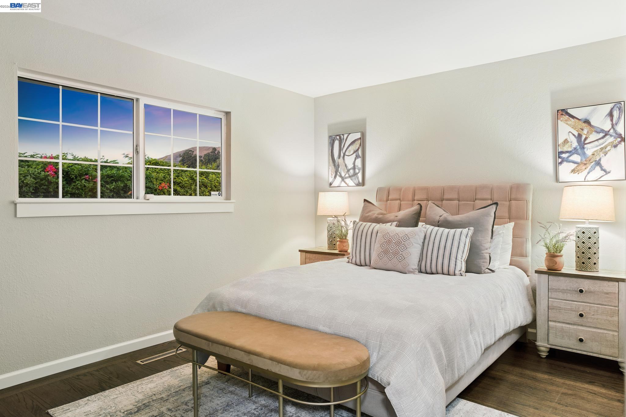 Staged bedroom with an upholstered bed, bench, and wide window overlooking flowering shrubs and a mountain.