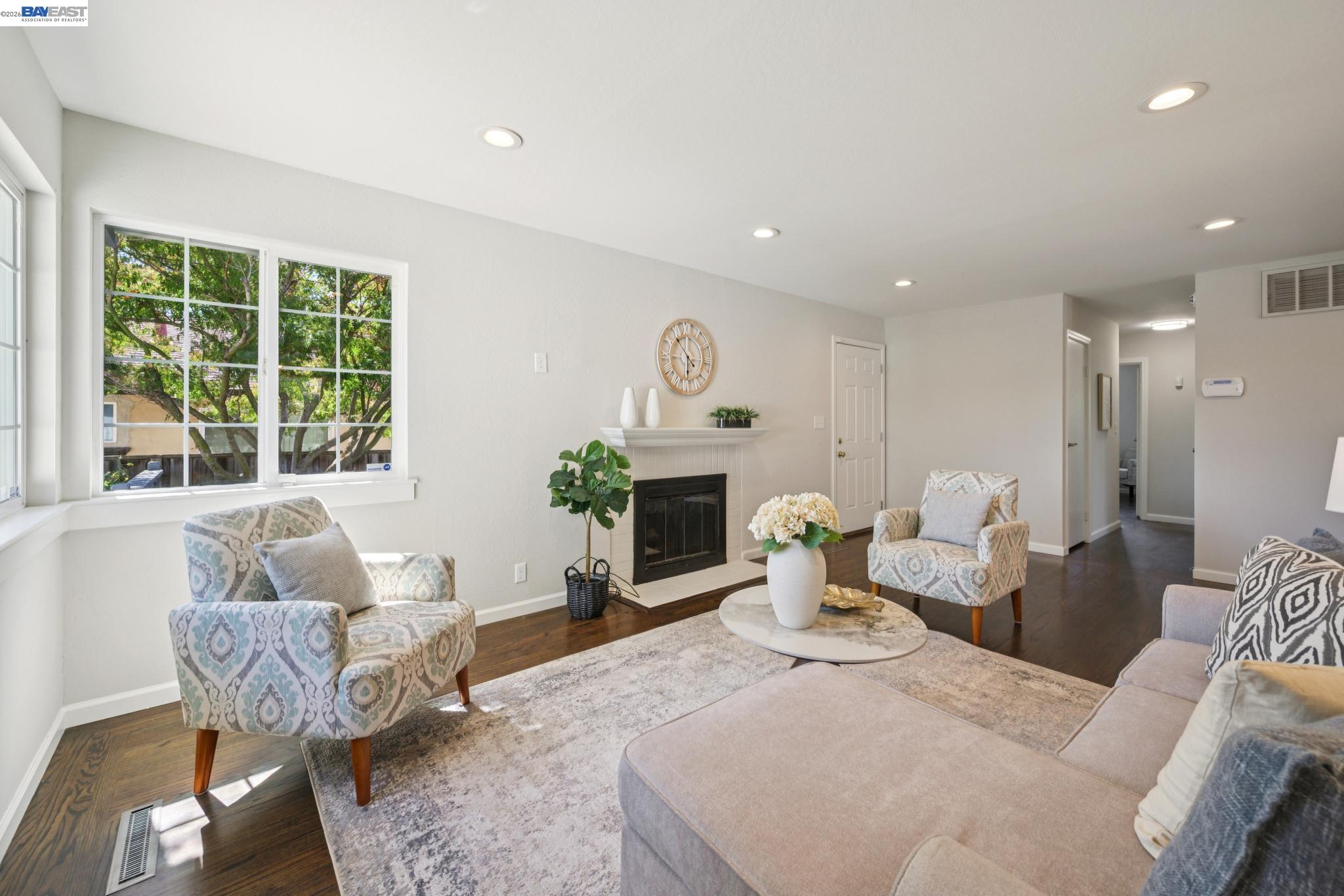 Staged living room with large leafy-window views, patterned armchairs, and a fireplace.