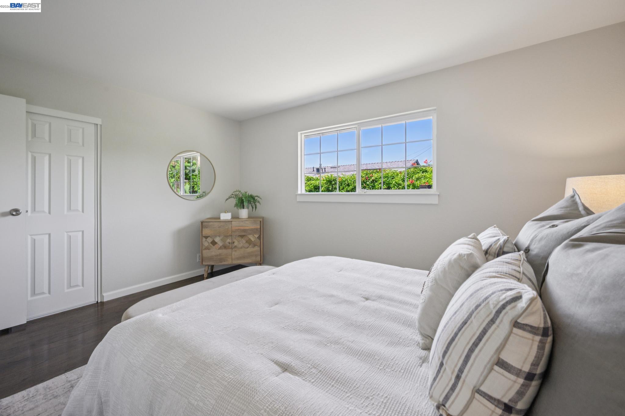 Bright bedroom with white bedding, a large window, and a small wooden cabinet.
