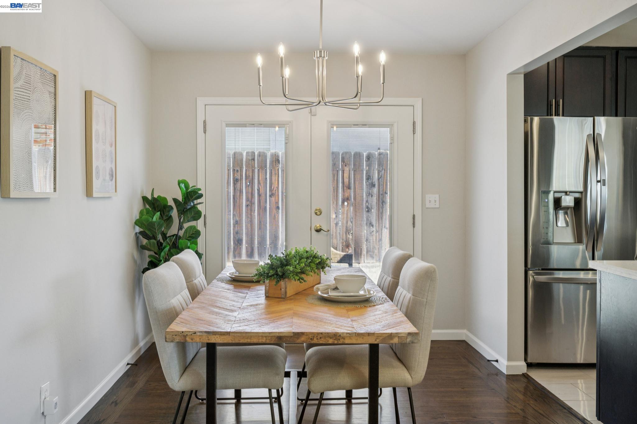 Dining area with a wood table and chandelier beside double glass doors and a stainless steel refrigerator.