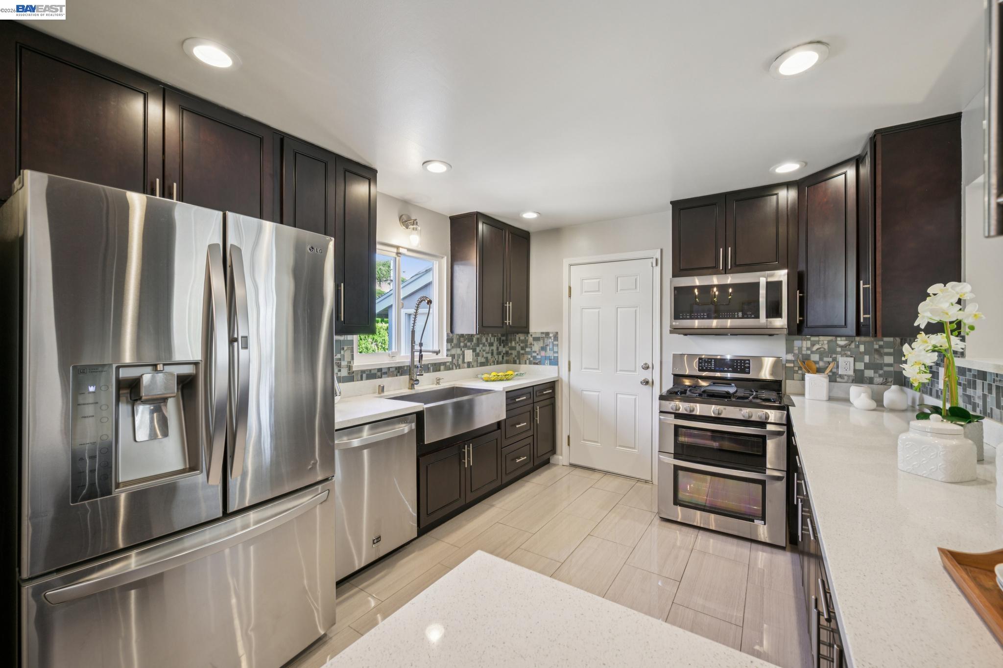 Kitchen with dark cabinets, white counters, stainless appliances, and a farmhouse sink beneath a window.