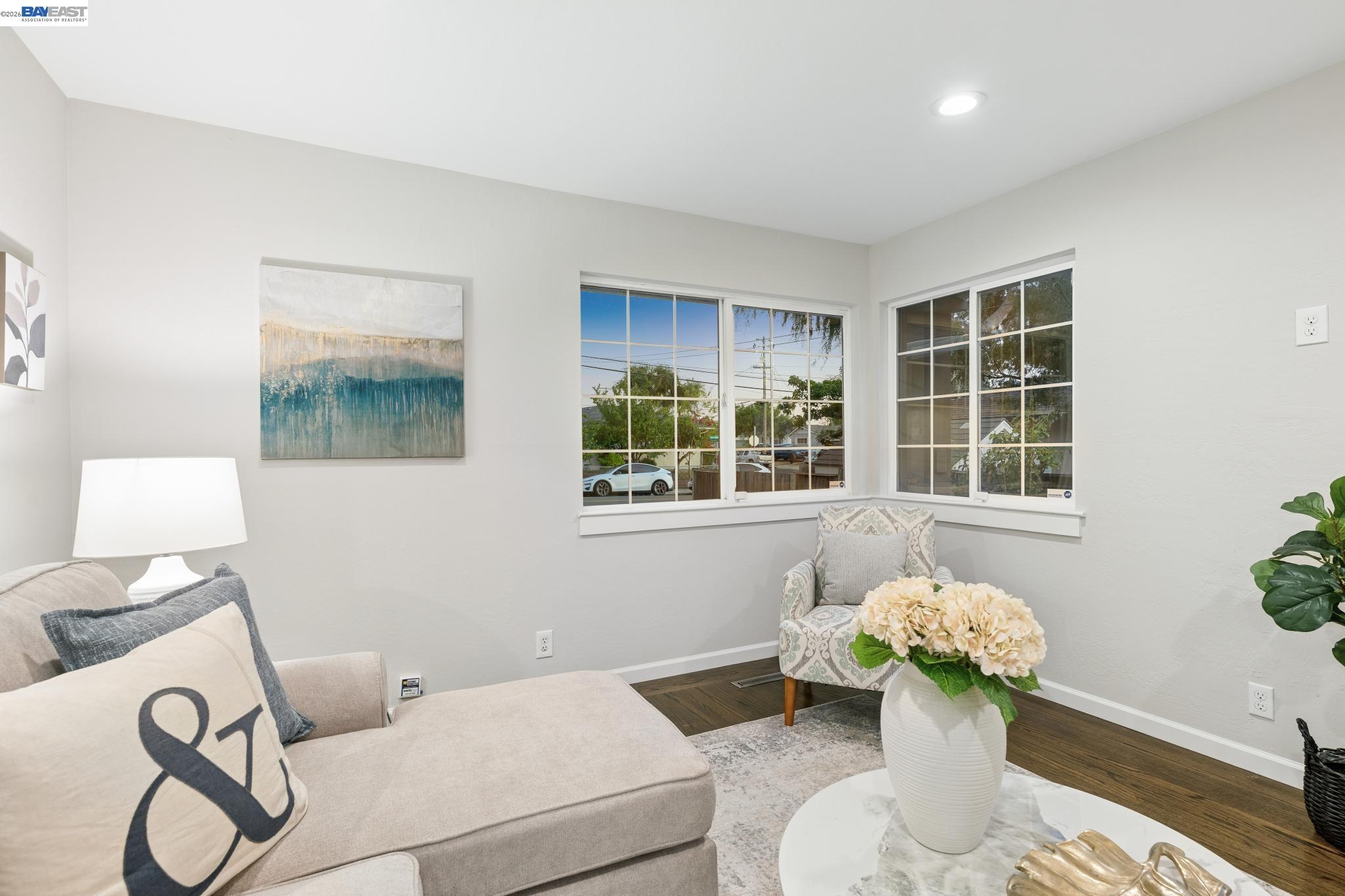 Staged living room with a beige sectional and corner windows overlooking a residential street.