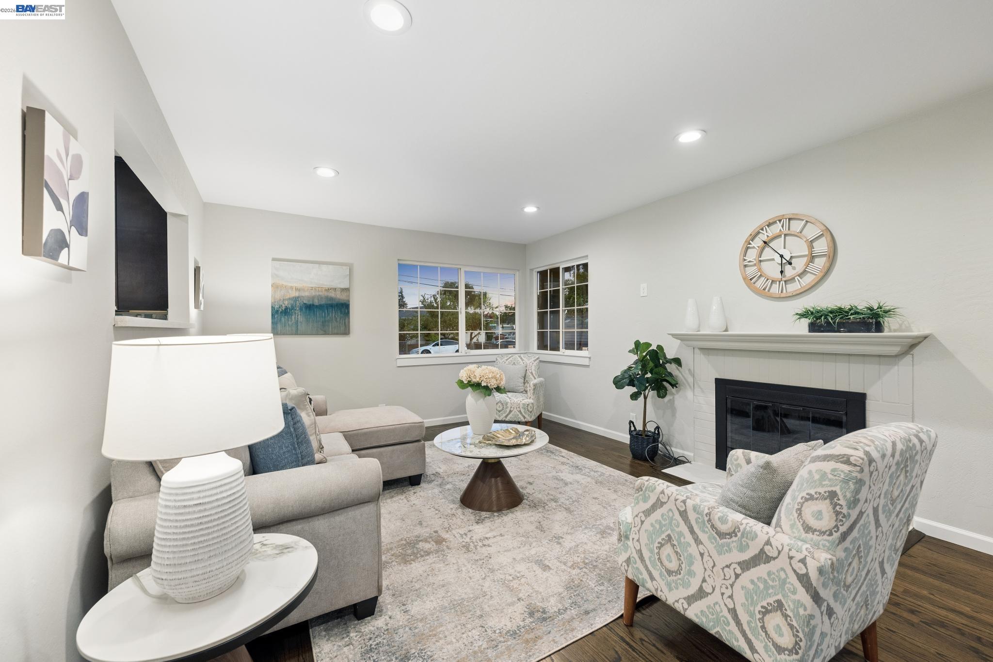 Staged living room with a white fireplace, gray sofa, patterned armchair, and corner windows.