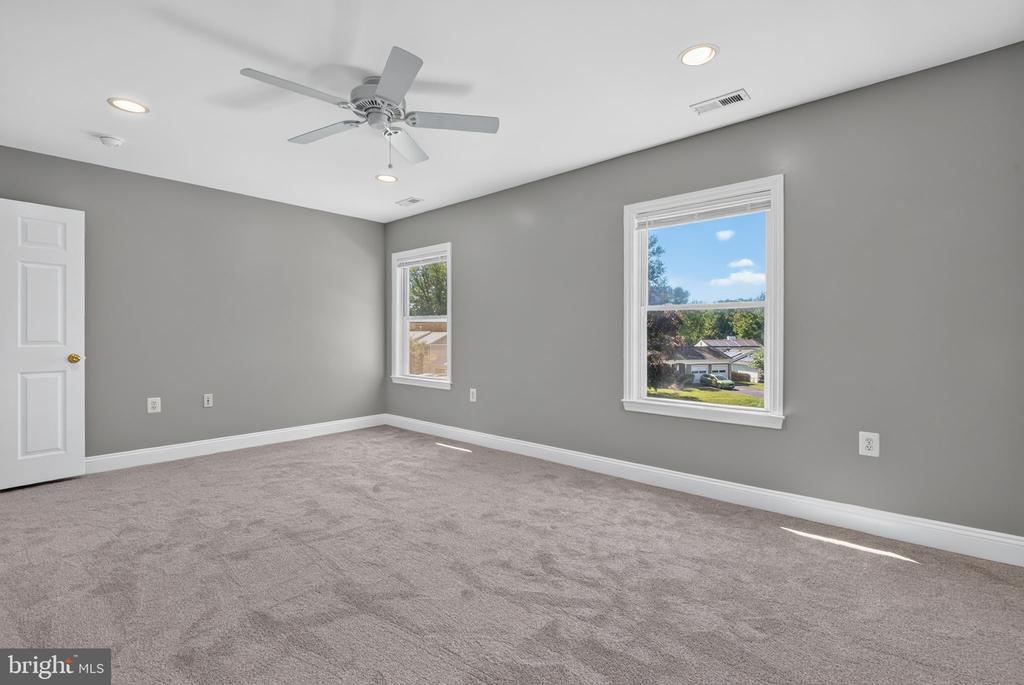 Empty bedroom with gray walls, taupe carpeting, two windows, and a ceiling fan.