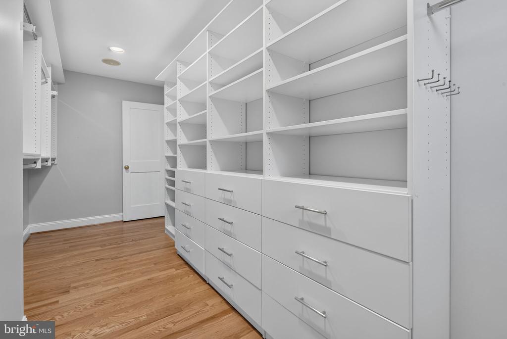 Walk-in closet with white built-in shelving, drawers, and hardwood flooring.