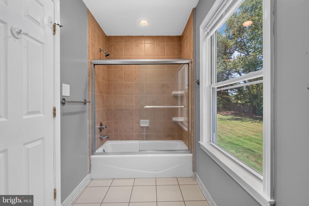 Bathroom with a glass-enclosed tub and tan tile beside a large window overlooking the yard.