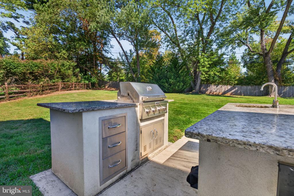 Outdoor kitchen with built-in grill and stone counters beside a fenced, tree-shaded lawn.