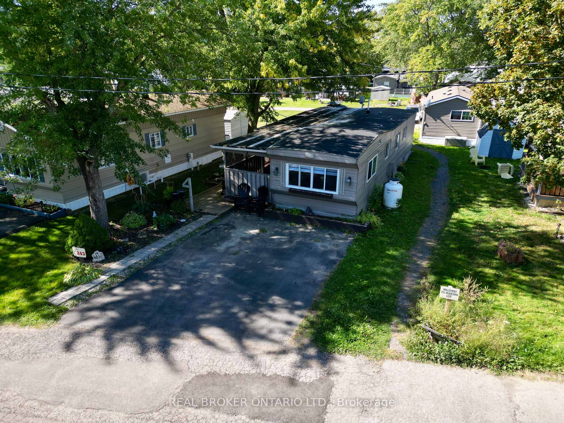 Gray manufactured home with a covered porch, paved parking area, and mature trees.